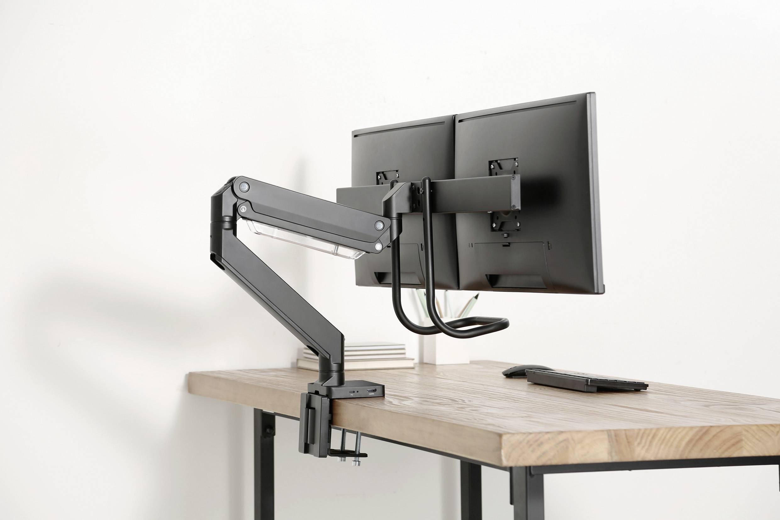 Ein Schreibtisch mit einem doppelten Monitorhalterungsarm, auf denen zwei Monitore befestigt sind. Der Tisch ist aus Holz, modern.