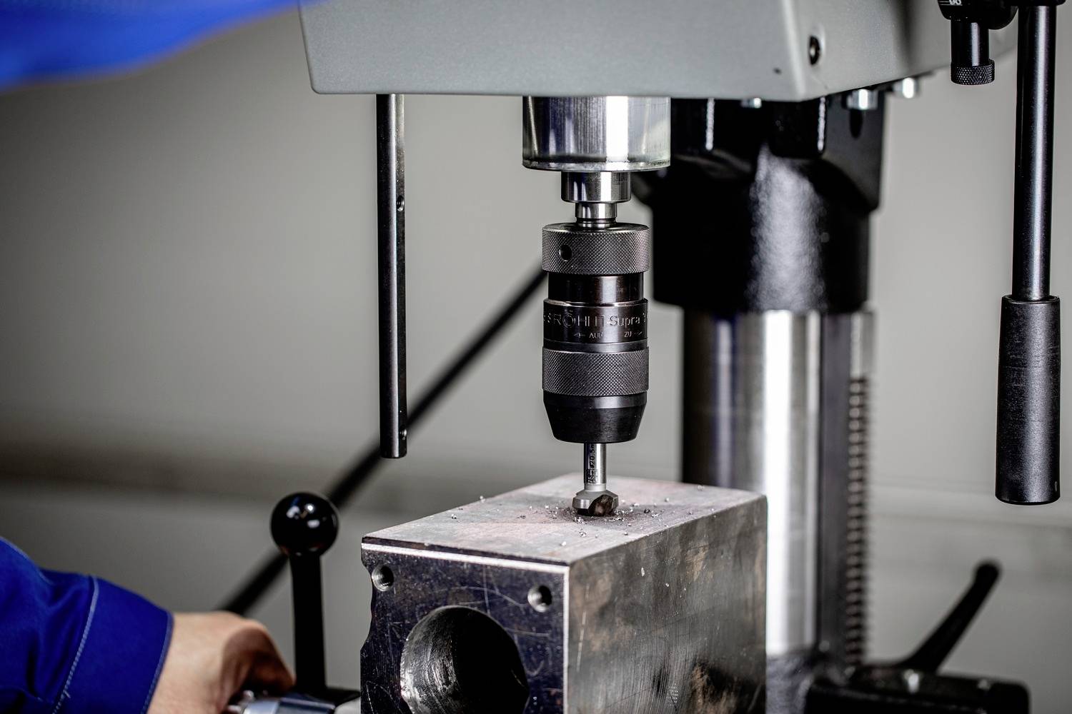 Eine Bohrmaschine bearbeitet einen Metallblock. Ein Arbeiter im blauen Overall bedient die Maschine in einer Werkstattumgebung.