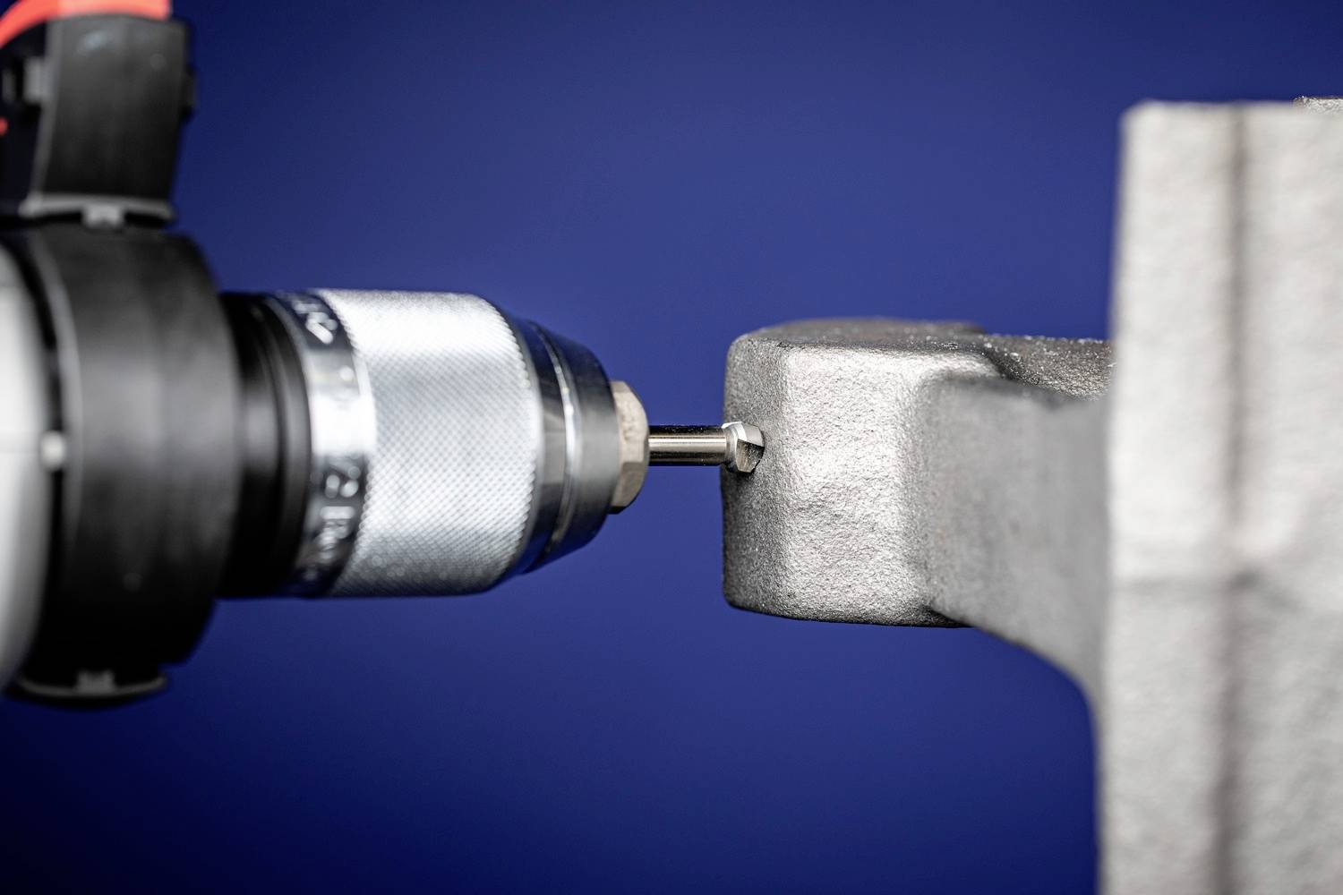 Ein Bohrer durchbohrt ein graues Metallstück vor einem blauen Hintergrund. Werkzeug ist im Fokus, zeigt handwerkliches Arbeiten.