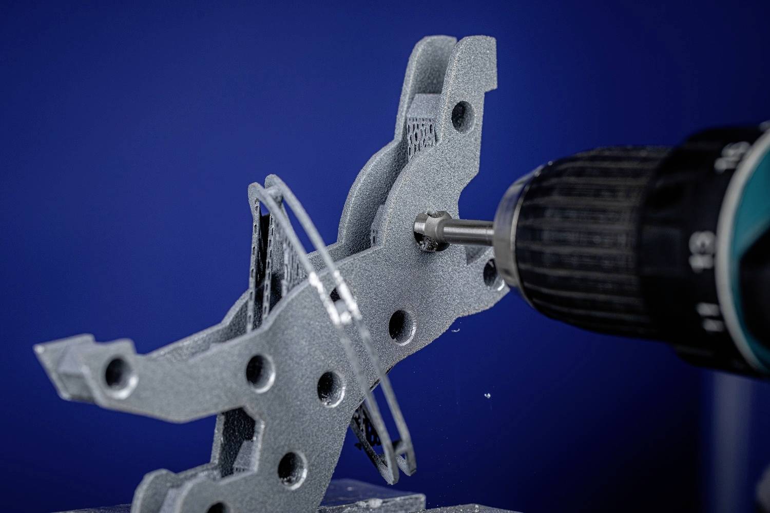 Eine Bohrmaschine bearbeitet ein graues Metallstück mit mehreren Löchern vor einem blauen Hintergrund. Funktion: Präzisionsarbeit.