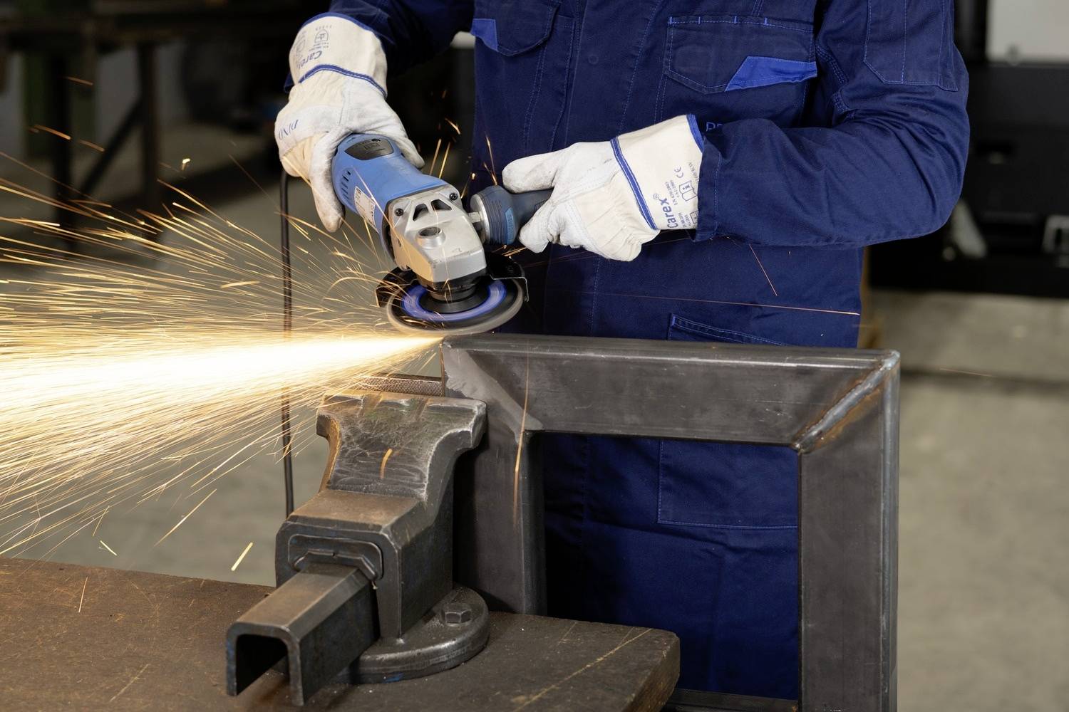 Person schleift Metall mit Winkelschleifer, Funken sprühen. Trägt blaue Schutzkleidung und weiße Handschuhe. Werkstattumgebung.