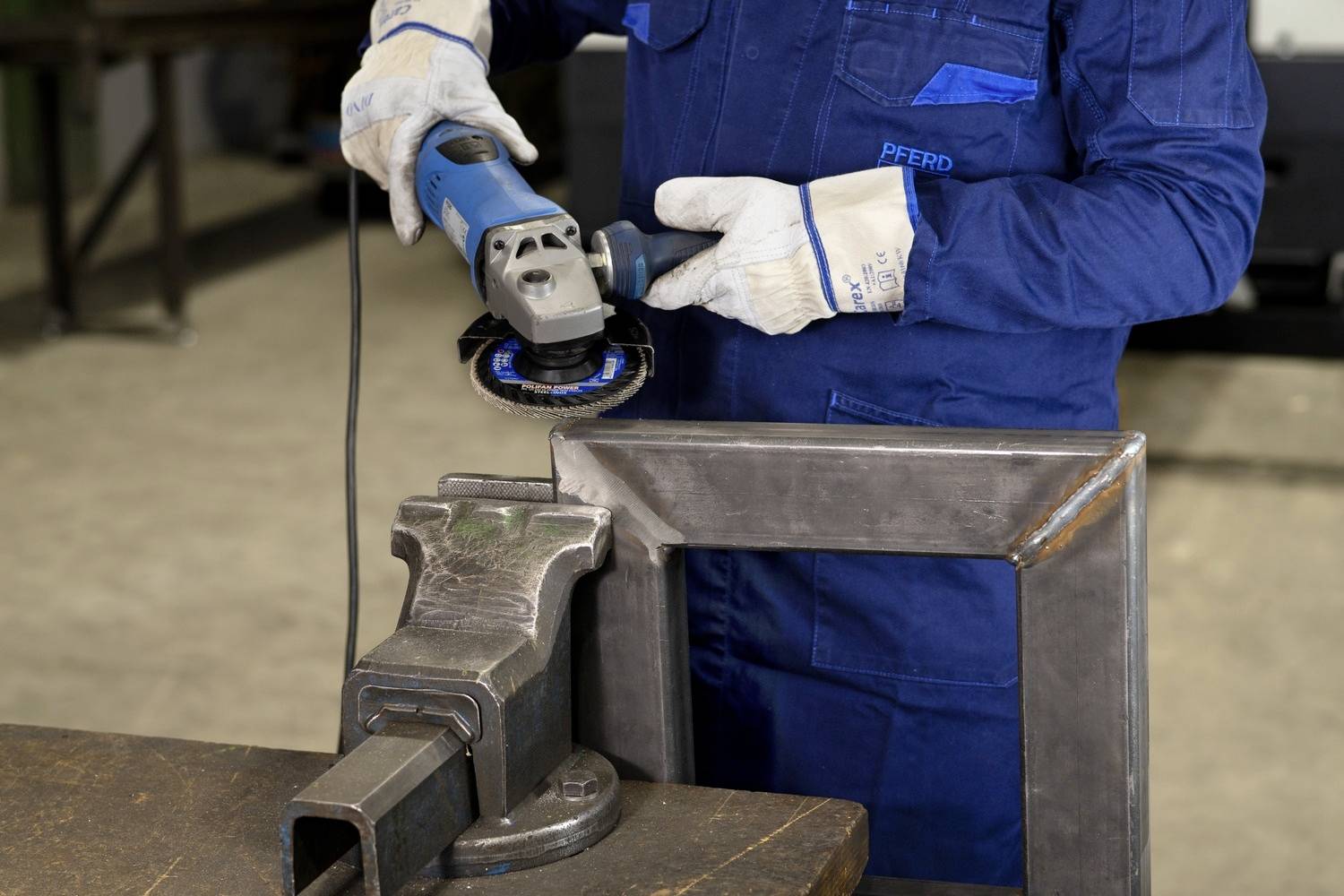 Ein Arbeiter in blauer Arbeitskleidung schleift mit einem Winkelschleifer ein Metallgestell, das in einem Schraubstock eingespannt ist.