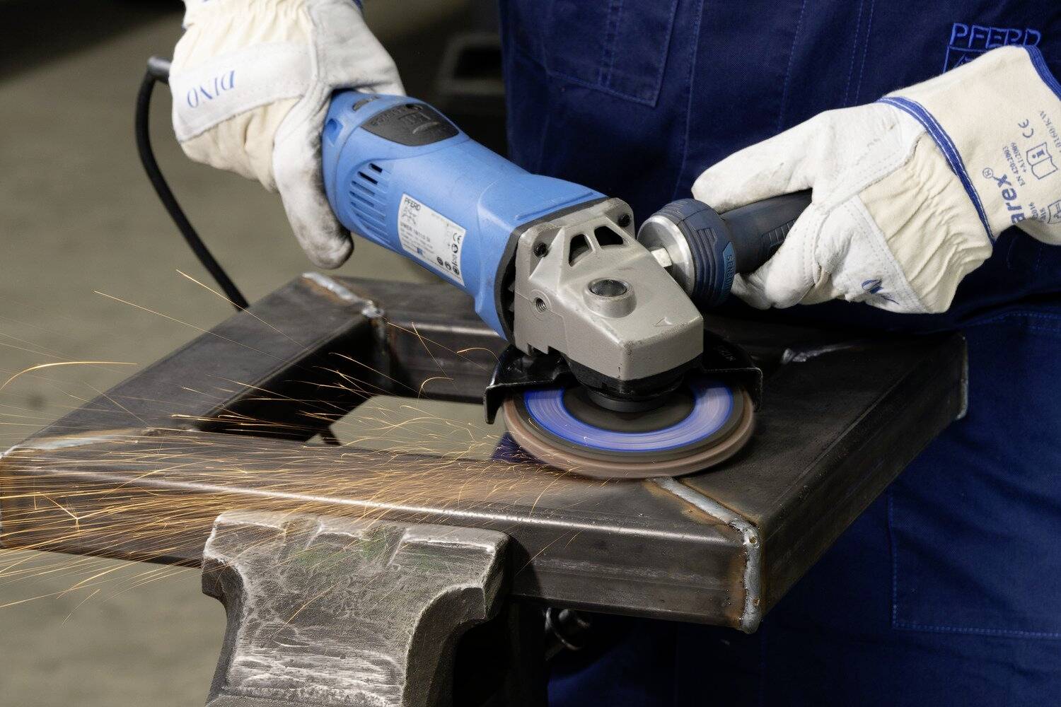 Eine Person schleift mit einem blauen Winkelschleifer ein Metallstück. Funken fliegen, während Schutzkleidung getragen wird.