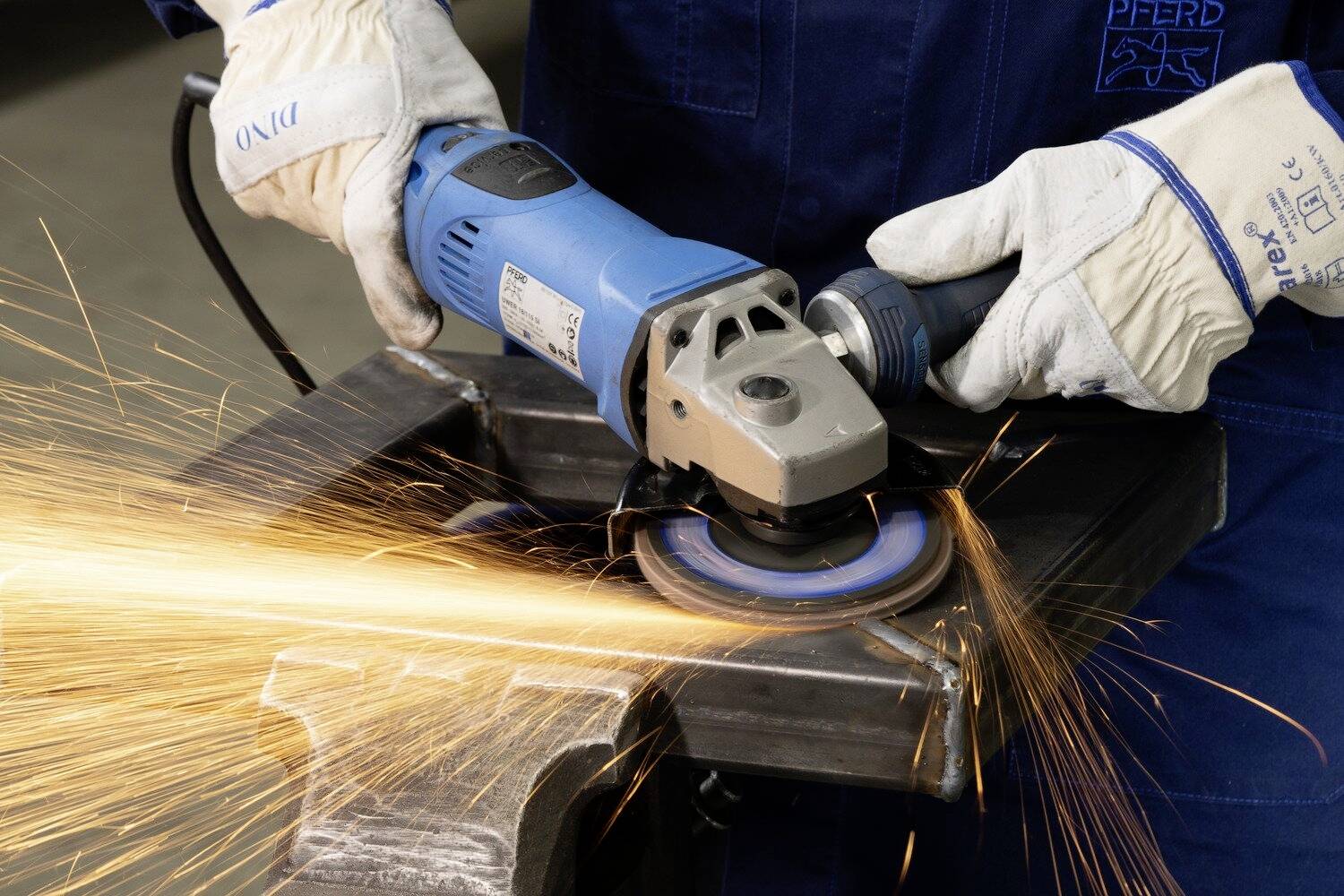 Person in blauer Arbeitskleidung schleift Metall mit einer Winkelschleifmaschine. Funken fliegen, Schutzkleidung wird getragen.