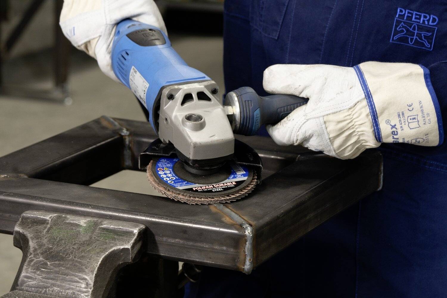 Eine Person schleift eine metallene Rahmenstruktur mit einem Winkelschleifer. Sie trägt Schutzhandschuhe und Arbeitskleidung.