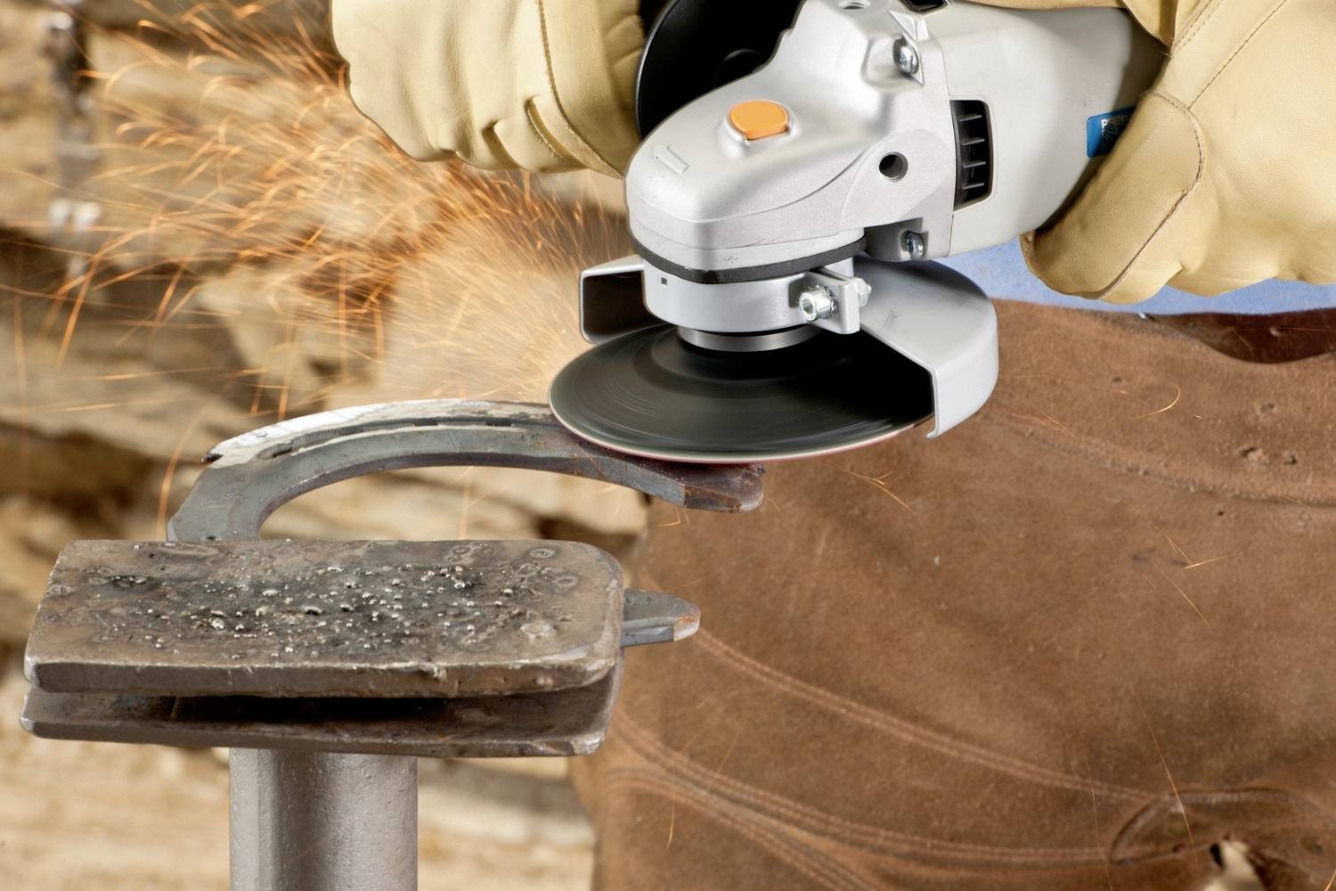 Person schleift mit Winkelschleifer ein Metallstück; Funken sprühen. Schutzhandschuhe und Lederkleidung werden getragen.