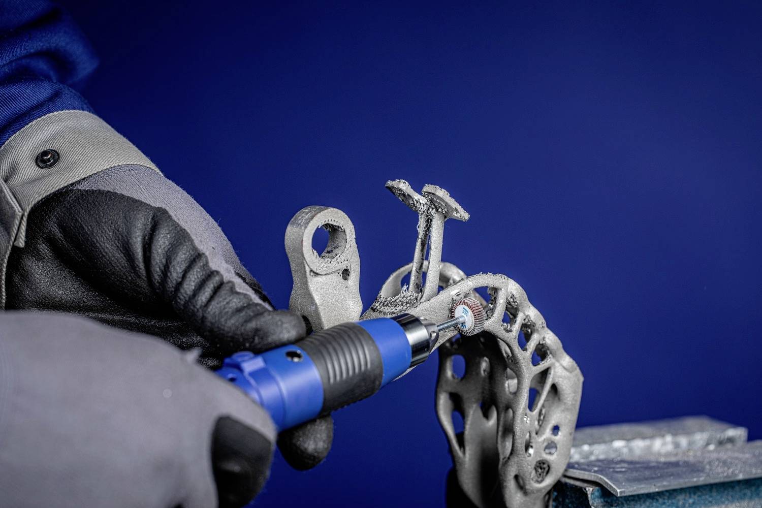 Eine Person in Schutzhandschuhen poliert ein komplex geformtes Metallteil mit einem blauen Handgerät vor blauem Hintergrund.