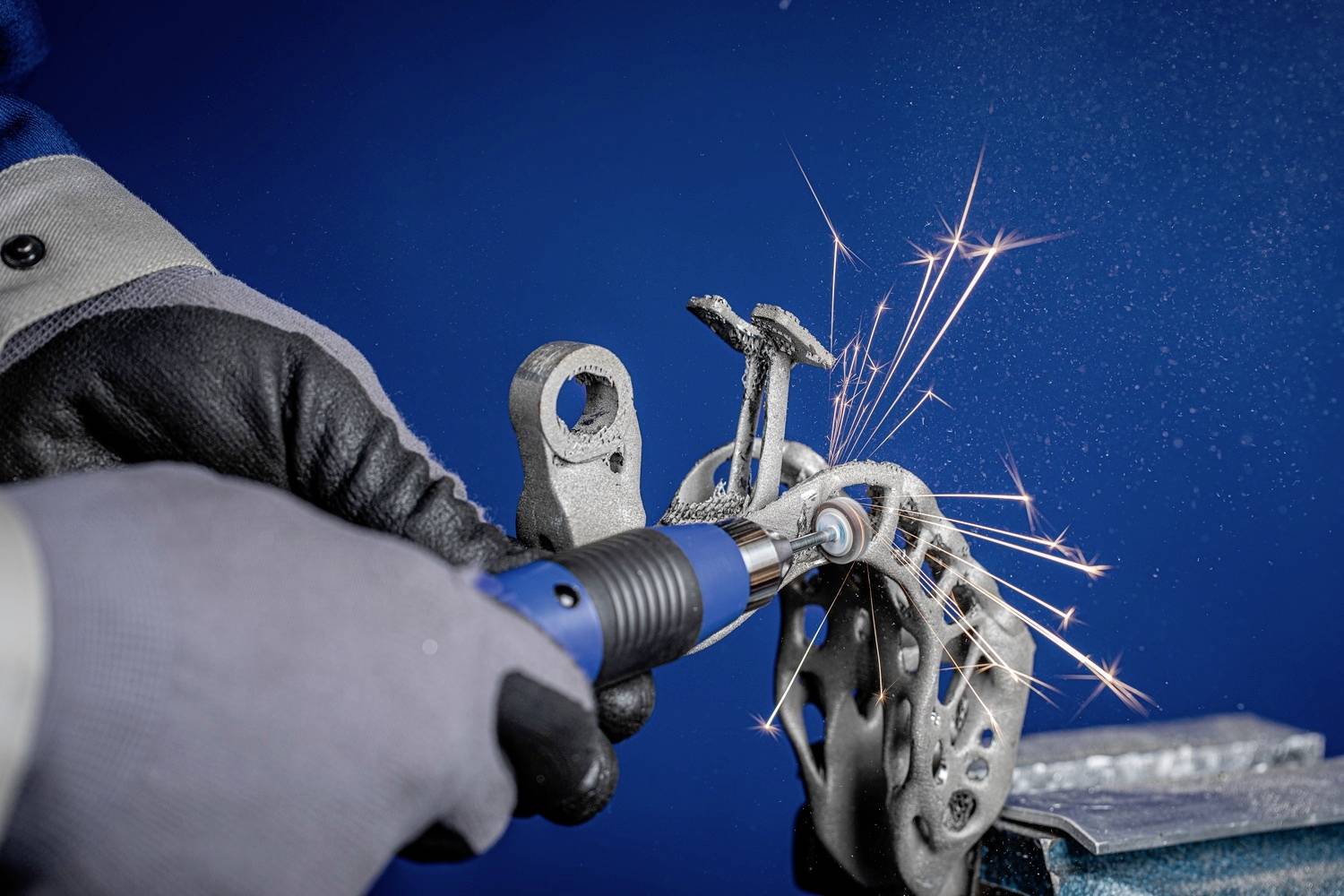 Eine Person schleift Metall mit einem Elektrowerkzeug, Funken sprühen. Sie trägt Handschuhe und Sicherheitskleidung vor einem blauen Hintergrund.