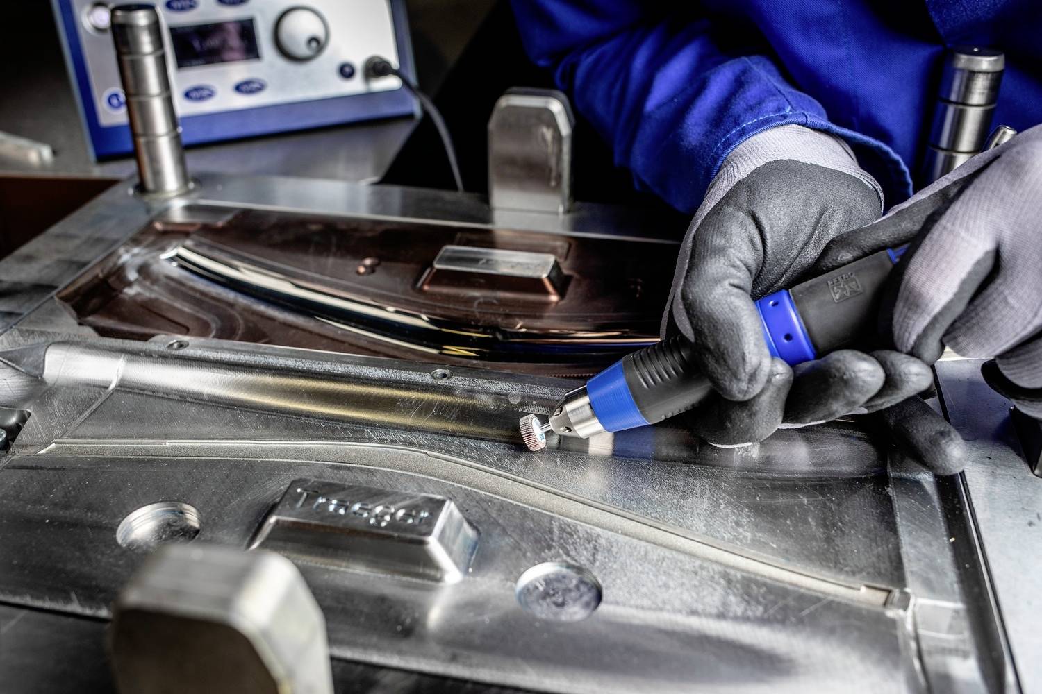 Person in Arbeitskleidung bearbeitet Metallform mit einem elektrischen Werkzeug. Im Hintergrund ein Kontrollgerät.