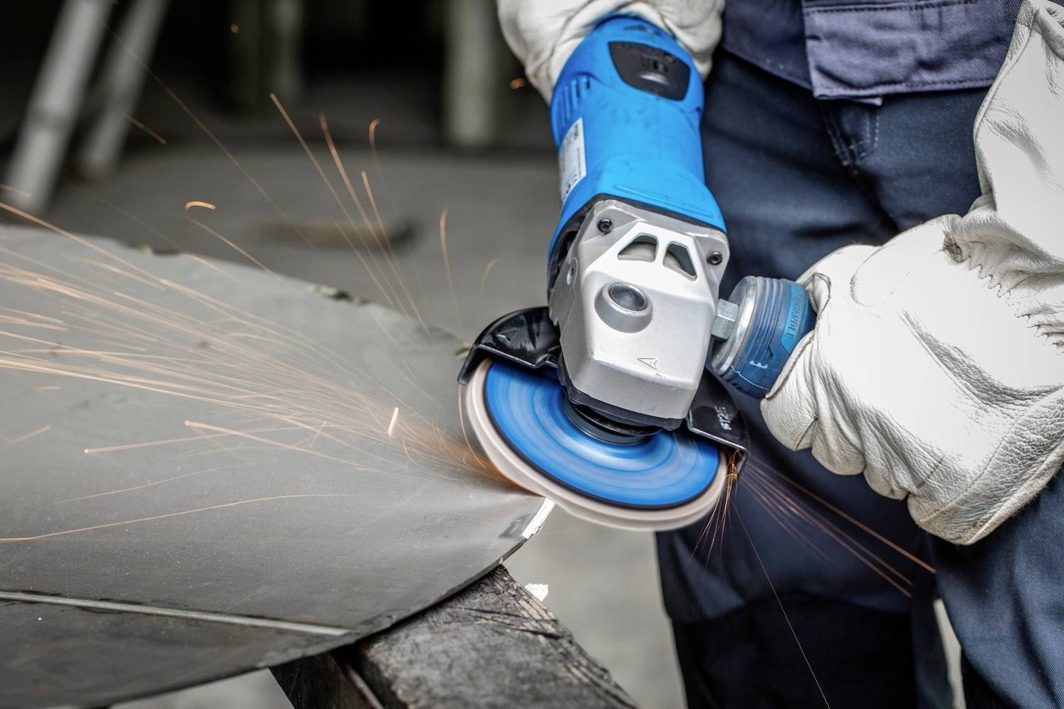 Person schleift mit einer blauen Winkelschleifmaschine Metall, Funken fliegen. Fokus auf Arbeitssicherheit mit Handschuhen.