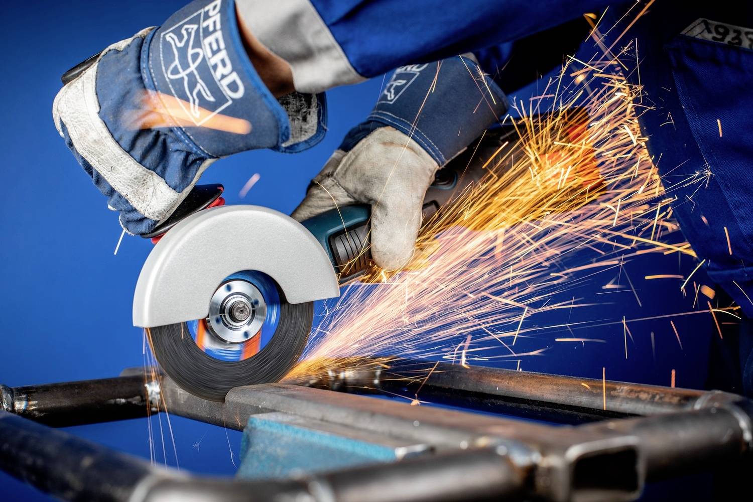 Handwerker schneidet mit Winkelschleifer Metallrohr. Funken fliegen in alle Richtungen. Arbeiter trägt Schutzhandschuhe und blaue Arbeitskleidung.