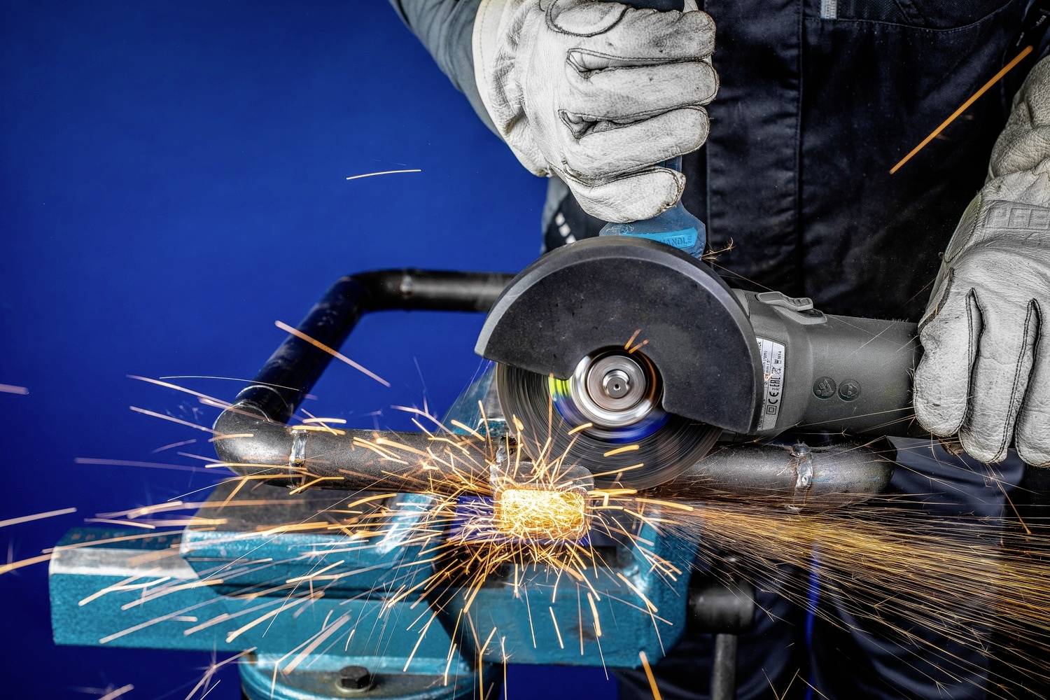 Eine Person schleift ein Metallrohr mit einem Winkelschleifer, Funken sprühen. Sie trägt Schutzhandschuhe und Arbeitskleidung.