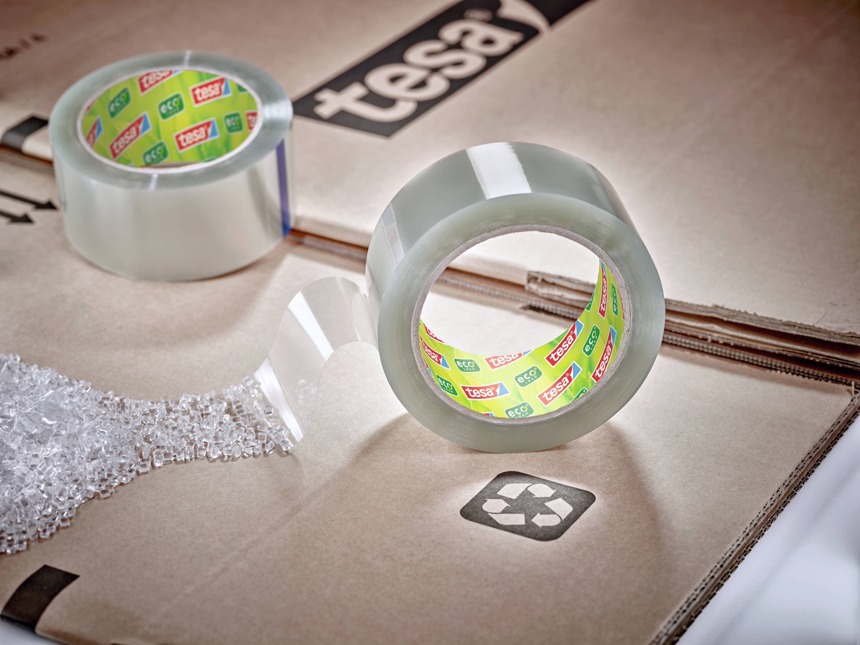 Eine Rolle durchsichtiges Klebeband liegt auf einem Karton mit Recycling-Symbol, daneben ist eine bereits geöffnete Rolle.