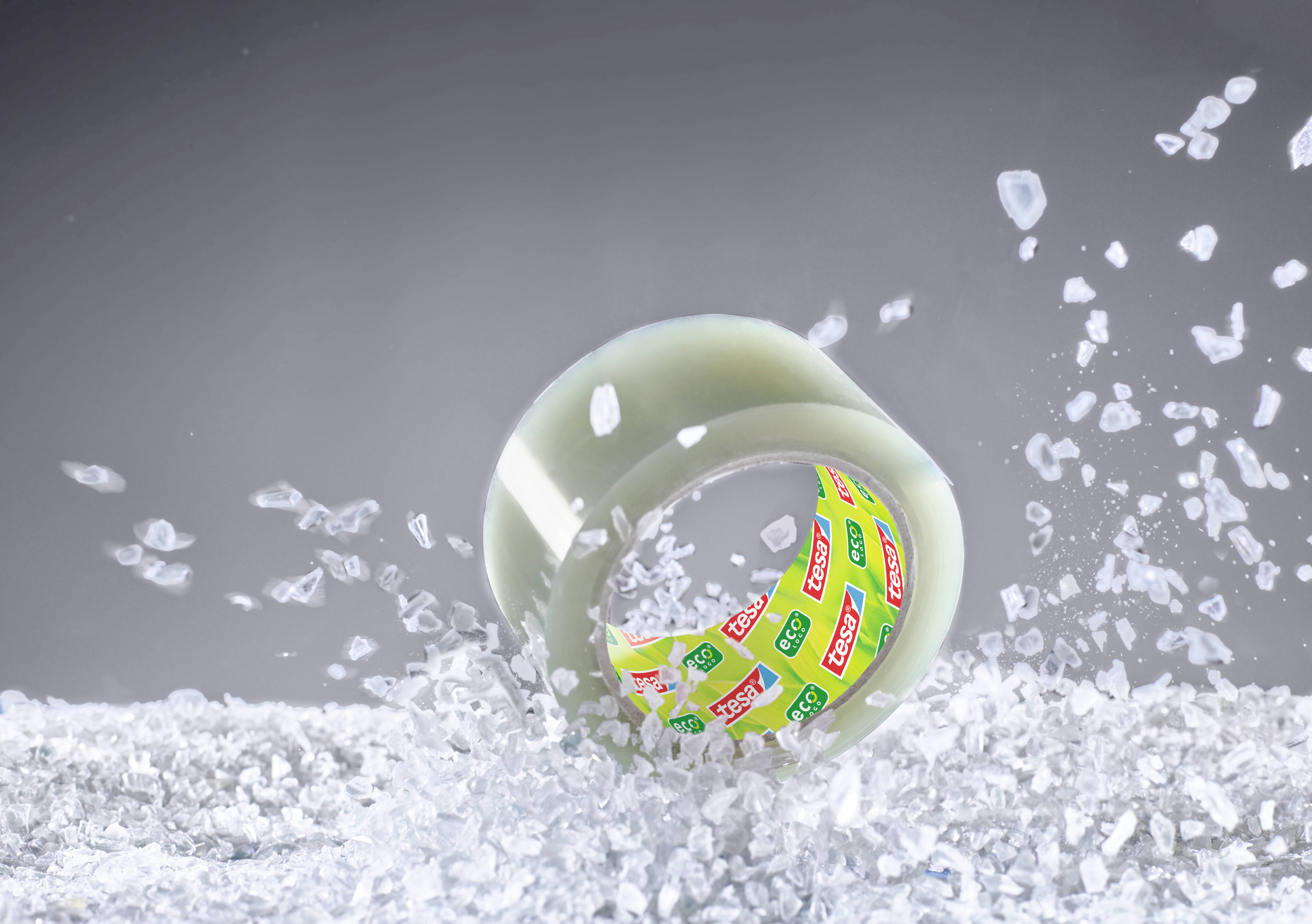 Eine Rolle transparentes Klebeband ist umgeben von zerbrochenem Glas in einer dynamischen Darstellung; Markenlogo sichtbar.