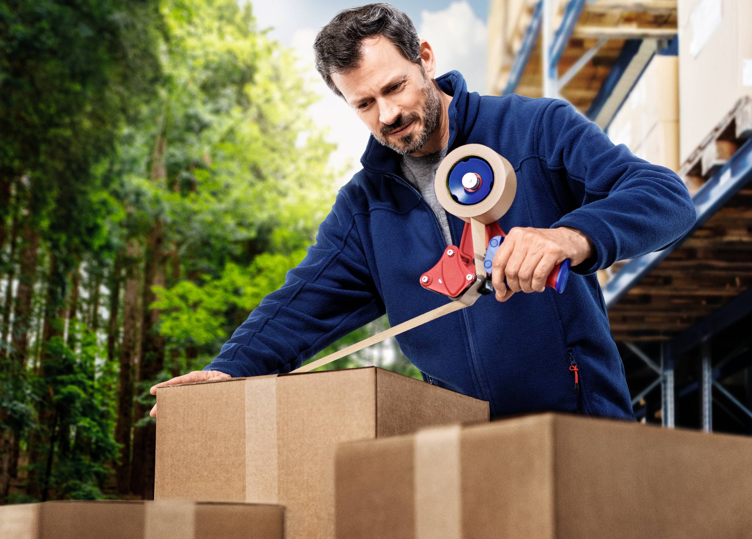 Ein Mann steht in einem Lagerhaus und verschließt mit Klebeband Kartons. Im Hintergrund sind Bäume und Regale sichtbar.