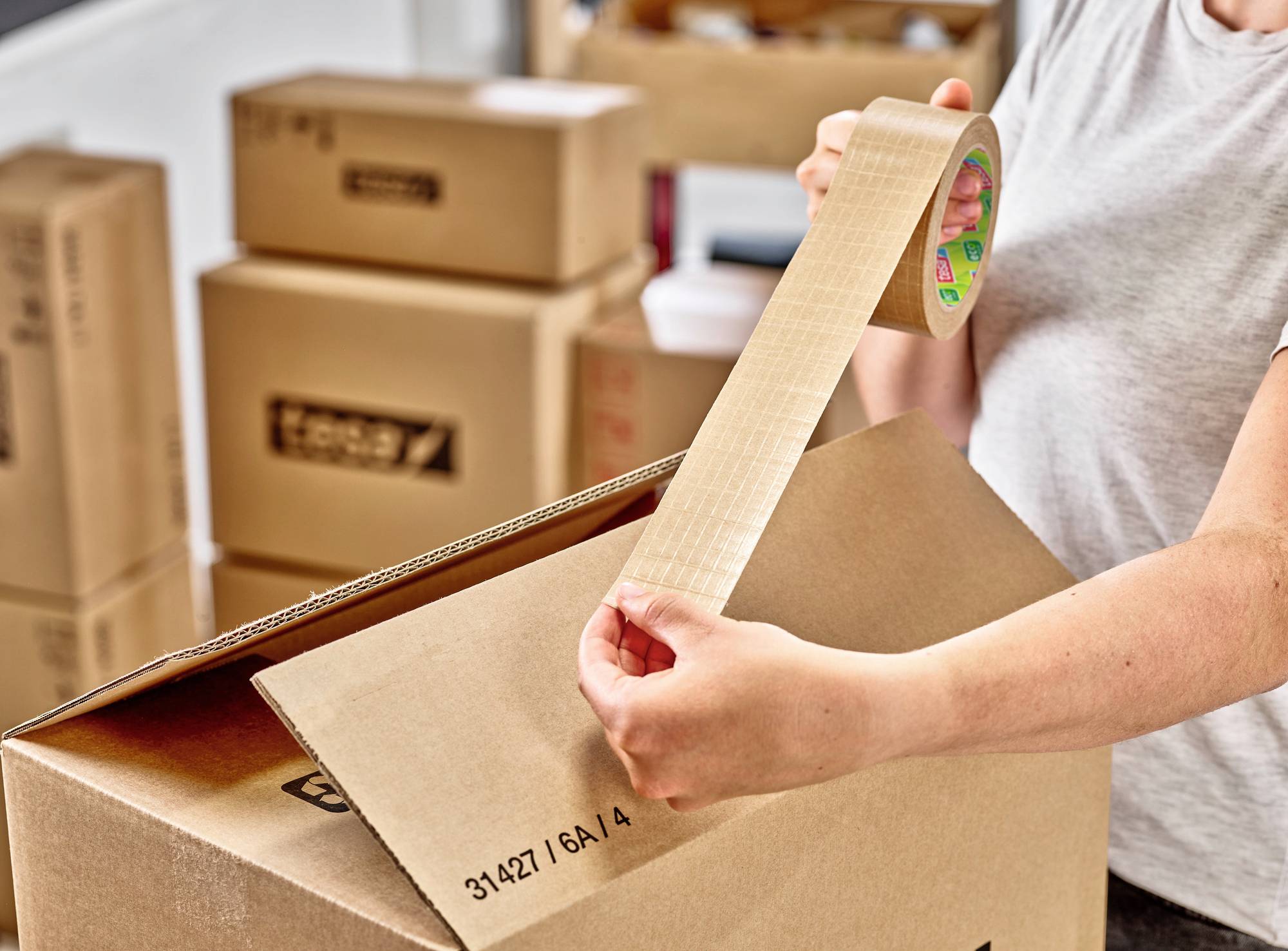 Eine Person mit einem weißen Shirt verschließt einen Karton mit Klebeband. Im Hintergrund sind weitere gestapelte Kartons sichtbar.