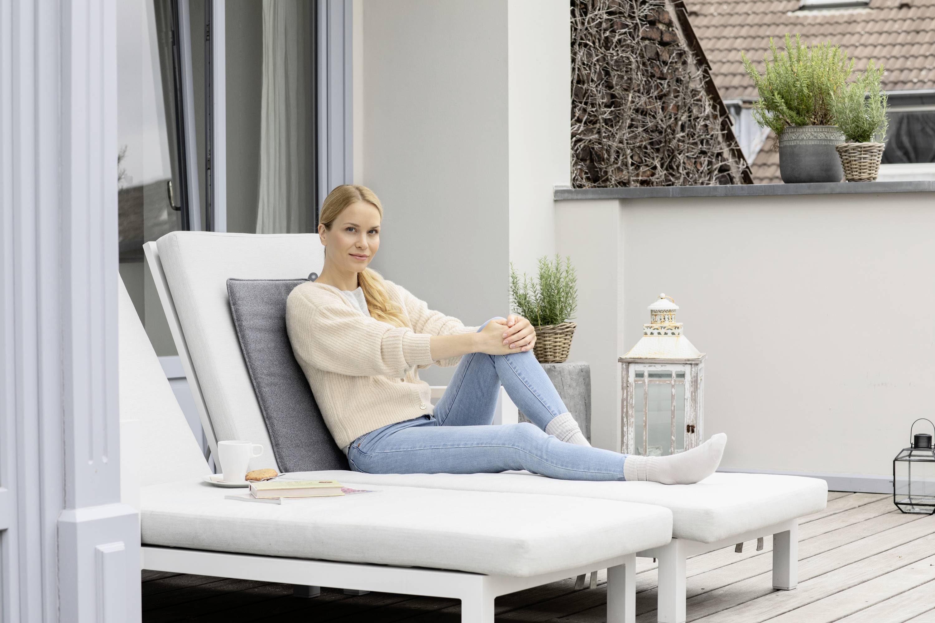 Eine Frau sitzt entspannt auf einer Terrasse auf einer weißen Liege, umgeben von Pflanzen und Dekorationen.