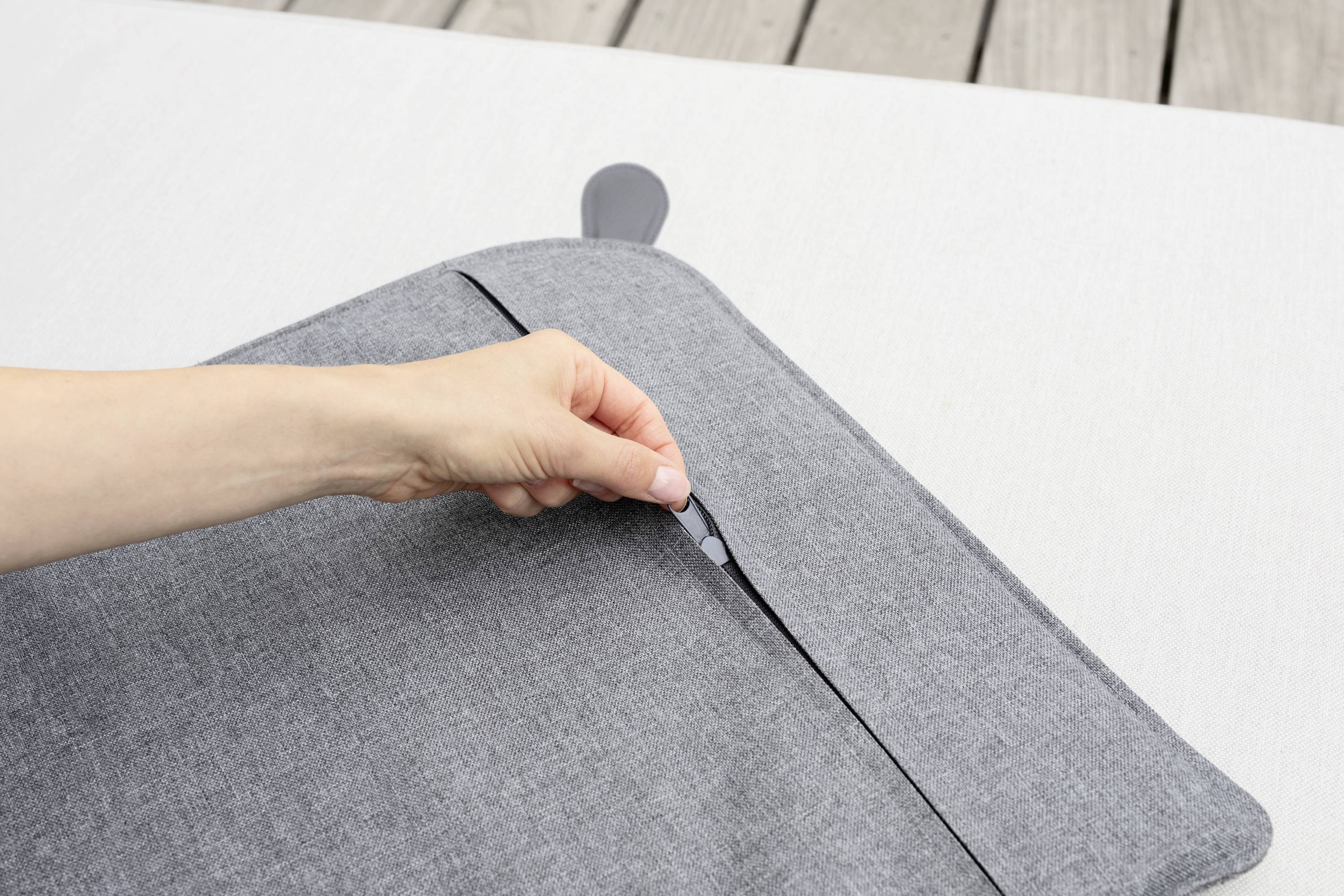Eine Hand öffnet einen grauen Reißverschluss auf einem Kissenbezug. Im Hintergrund ist ein heller Stoff sichtbar.