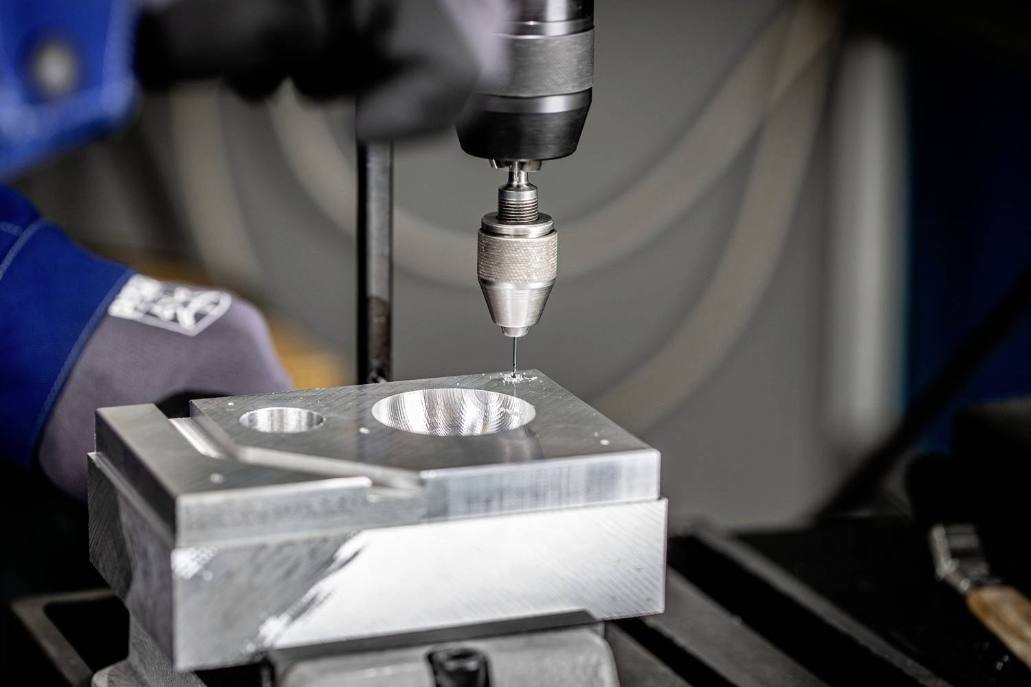 Eine Person bohrt mit einer Bohrmaschine ein Loch in ein Metallstück, das auf einer Werkbank liegt. Handschuhe schützen die Hände.