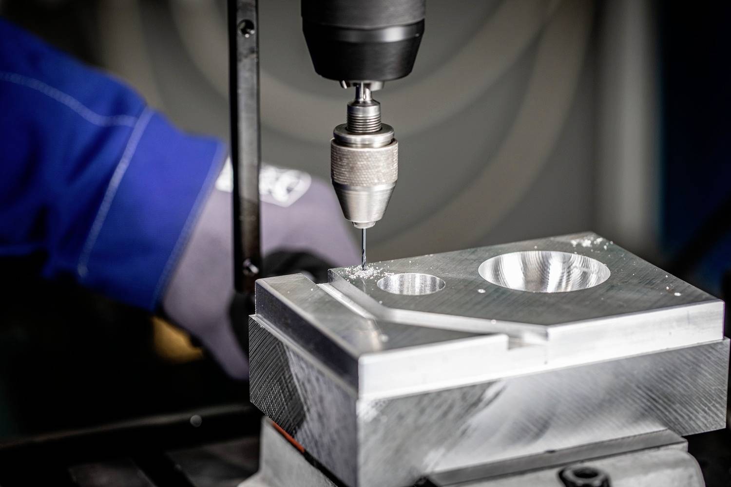 Mann in blauer Jacke und Handschuhen bedient eine Metallbohrmaschine, die präzise Löcher in ein silbernes Metallstück bohrt.