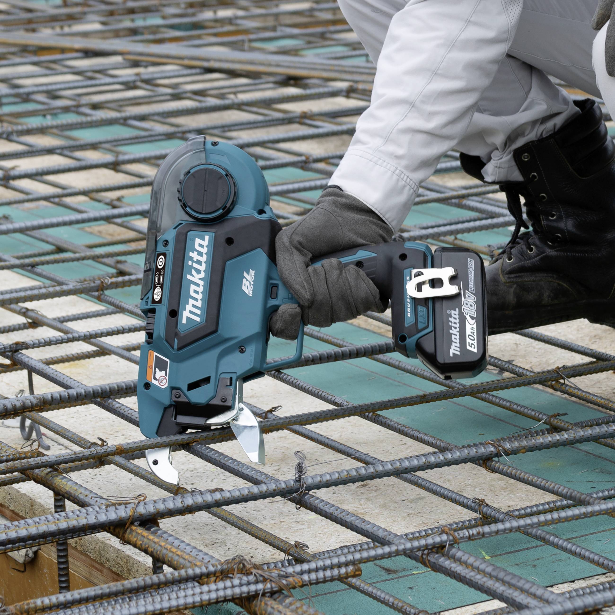 Eine Person befestigt Stahlbewehrungen auf einer Baustelle mit einem blauen Akku-Gerät. Sie trägt Arbeitshandschuhe und weiße Arbeitskleidung.