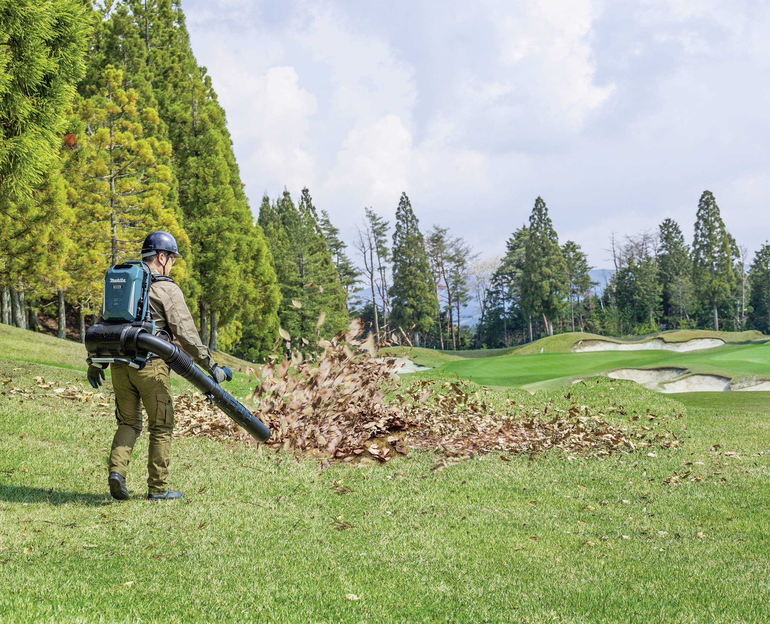 Eine Person bläst mit einem Laubbläser Blätter von einer Wiese. Im Hintergrund sind Bäume und ein Golfplatz zu sehen.