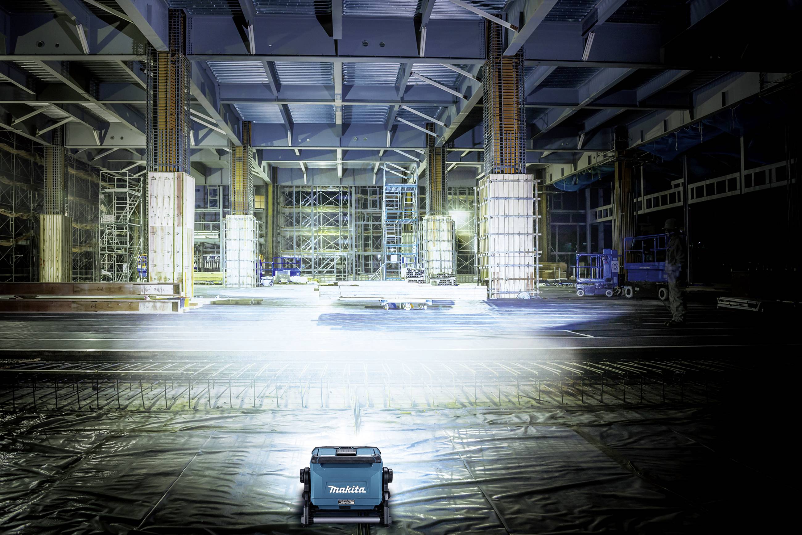 Ein großer, leerer Innenraum mit hohen Decken und Stahlträgern, beleuchtet von Bauleuchten. Im Vordergrund steht ein kleines, blaues Gerät.