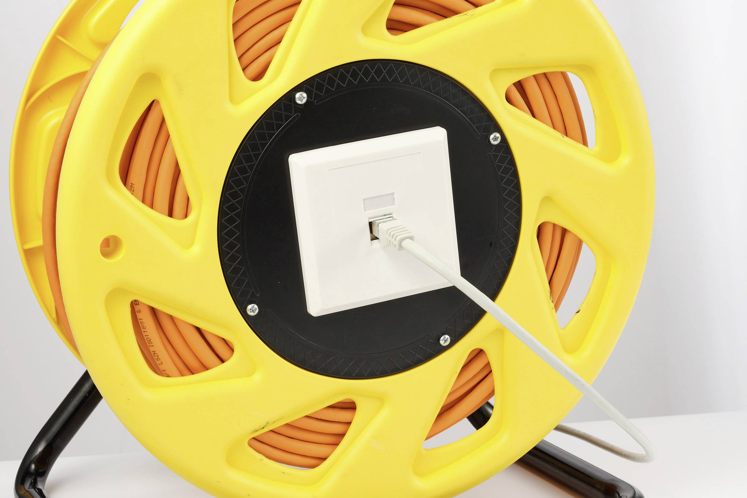 Gelbe Kabeltrommel mit aufgewickeltem orangefarbenem Kabel, LAN-Kabel durch Wandsteckdose vorne, auf schwarzem Ständer.