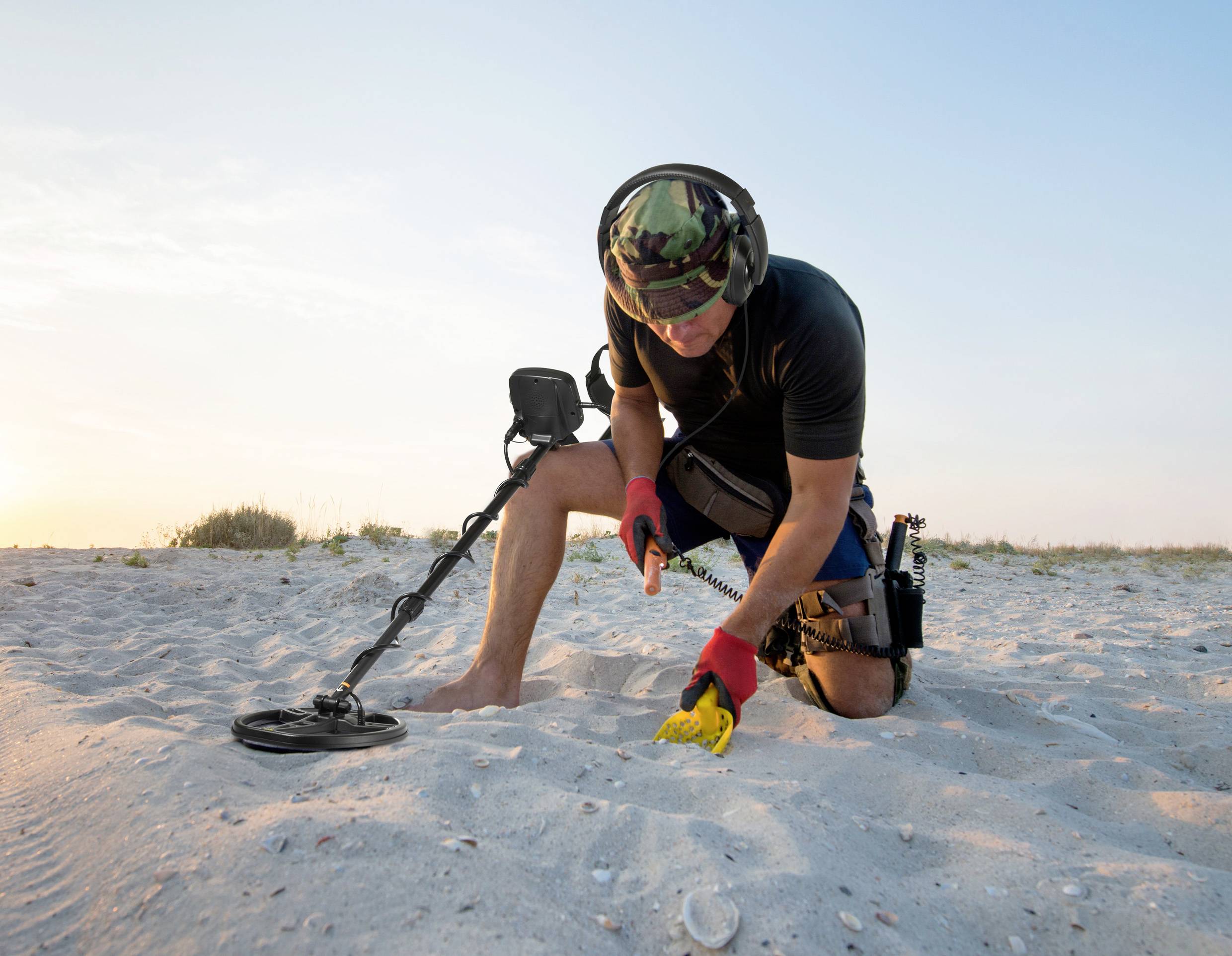 Person mit Metalldetektor kniet am Strand und untersucht den Sand. Es ist sonnig. Im Hintergrund sind Gräser und blauer Himmel zu sehen.