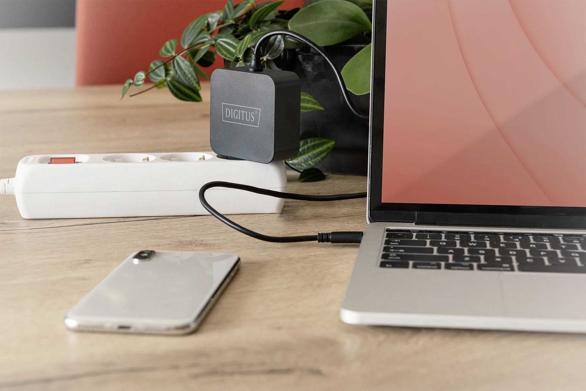Ein Laptop auf einem Tisch ist über ein USB-Kabel mit einem Ladegerät verbunden. Daneben liegen ein Smartphone und eine Steckdosenleiste.