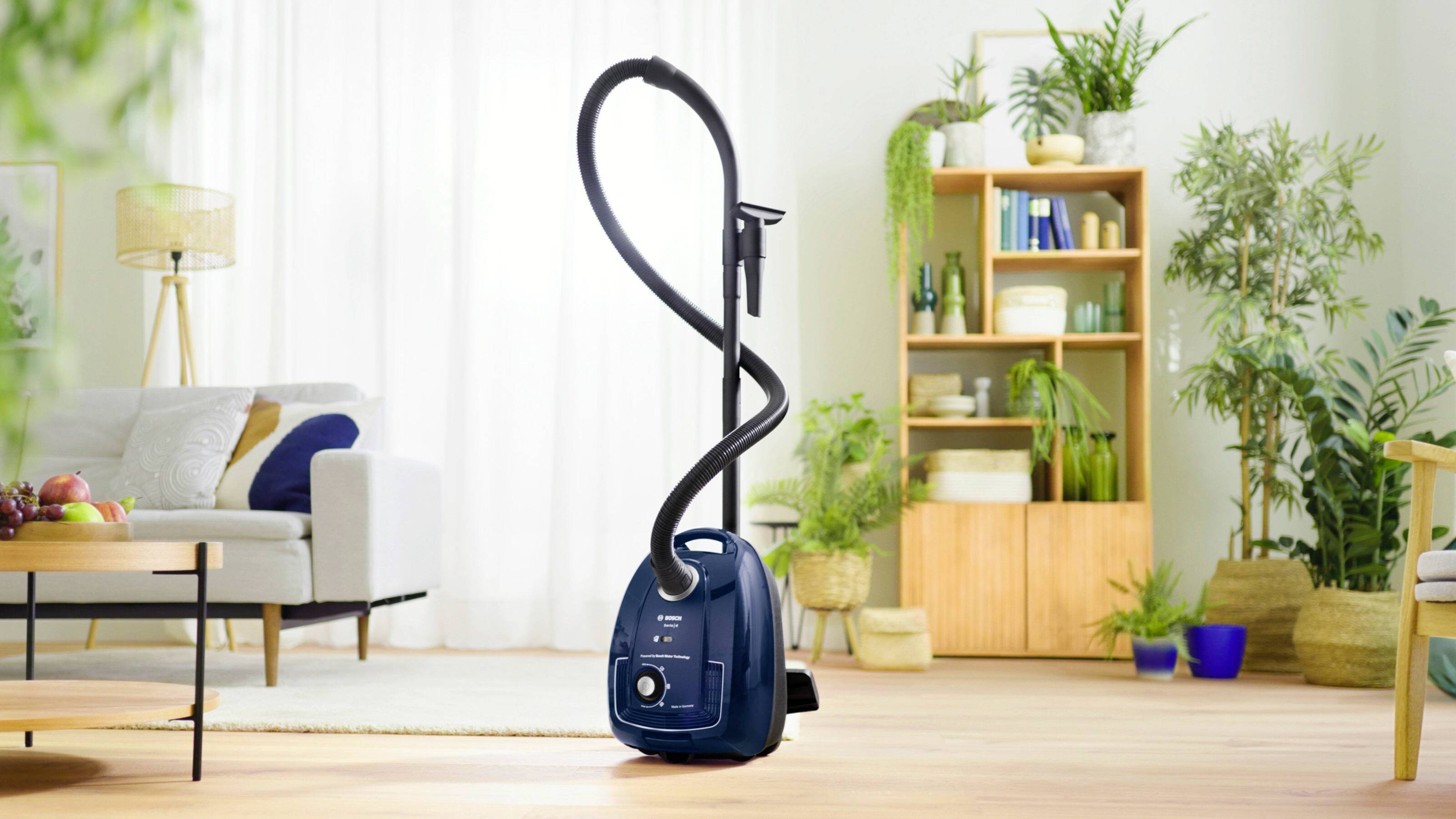 Ein Staubsauger steht mittig in einem hellen Wohnzimmer mit Pflanzen, Sofa und Regal im Hintergrund, bereit zum Einsatz.