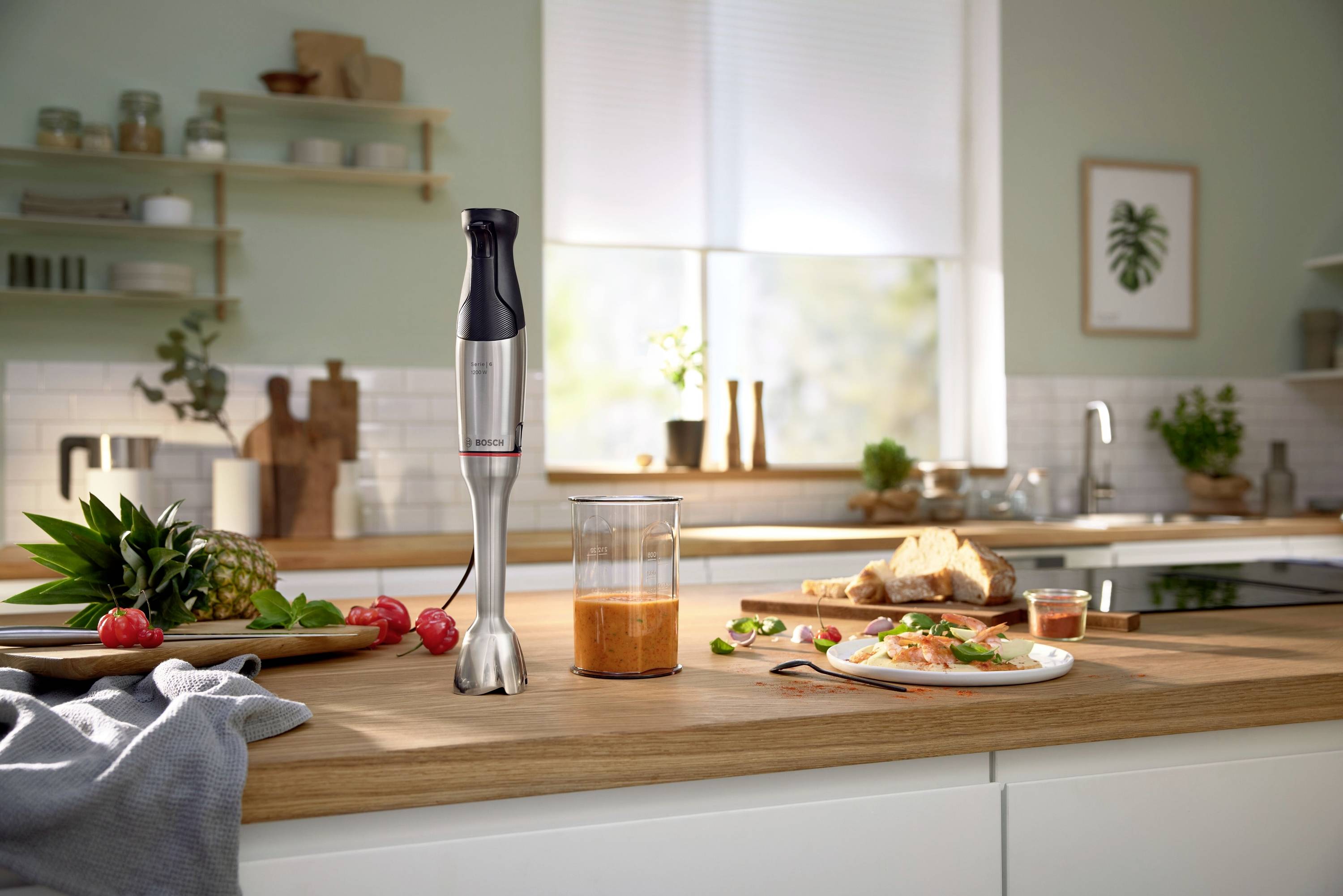 Ein Stabmixer steht auf einer Küchenarbeitsplatte neben frischen Früchten und einem Glas mit Smoothie. Im Hintergrund ist eine moderne Küche.