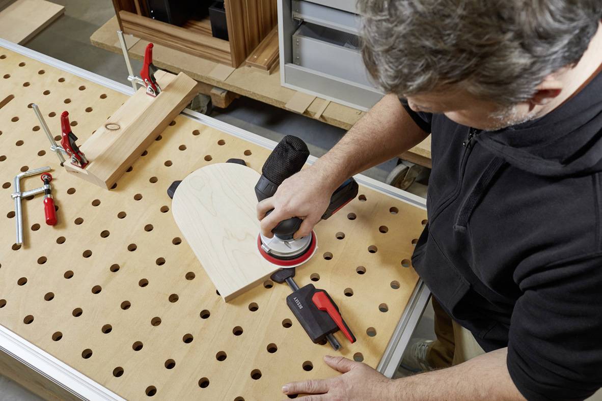Ein Mann bearbeitet ein herzförmiges Holzstück auf einem Lochbrett mit einer Schleifmaschine, umgeben von Werkzeugen.