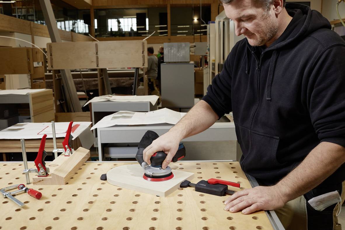 Ein Mann schleift in einer Werkstatt ein Holzstück auf einem Arbeitstisch. Umgeben von Werkzeugen und anderen Holzarbeiten.