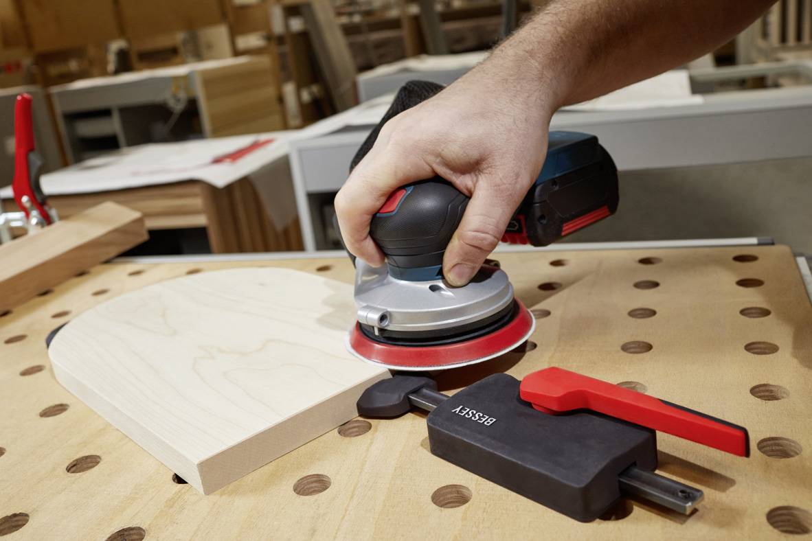 Eine Person schleift ein Holzstück auf einem Tisch mit einer rotierenden Schleifmaschine, umgeben von weiteren Werkstattgegenständen.