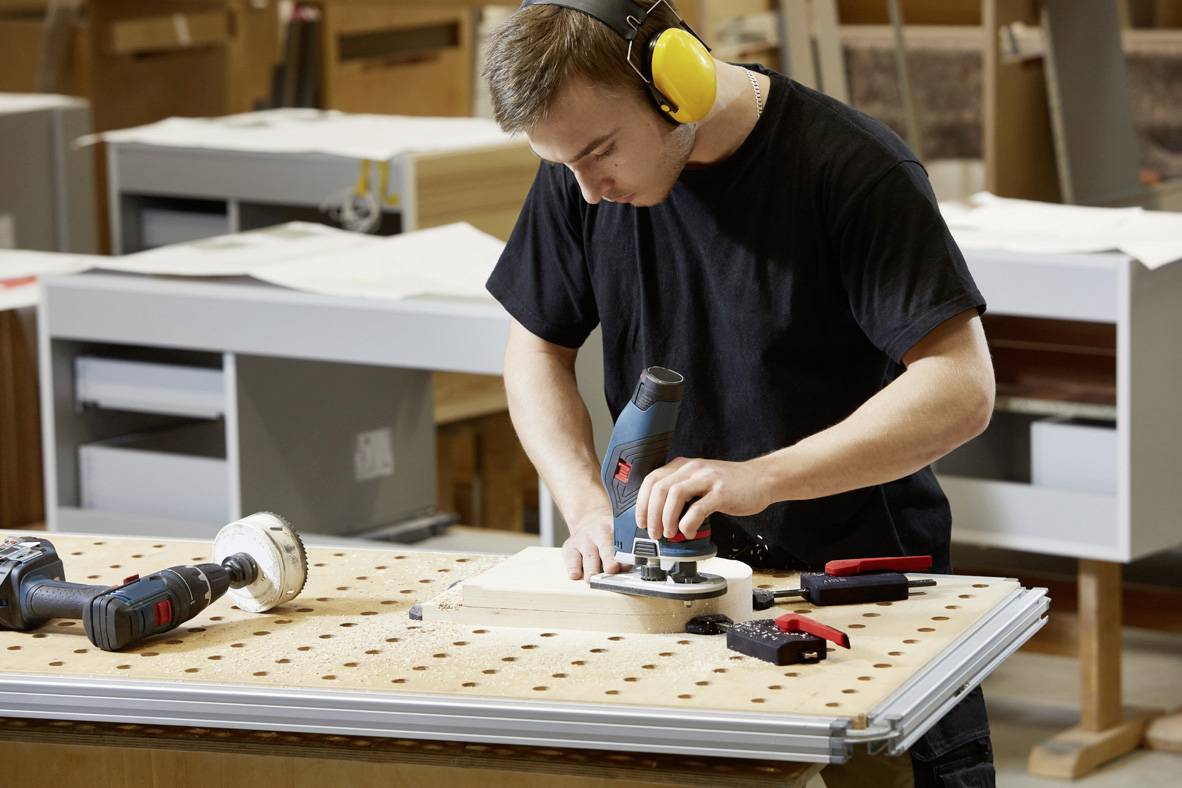 Ein Mann arbeitet mit einer Fräse an einem Holzbrett in einer Werkstatt. Er trägt Gehörschutz und ist von Werkzeugen umgeben.