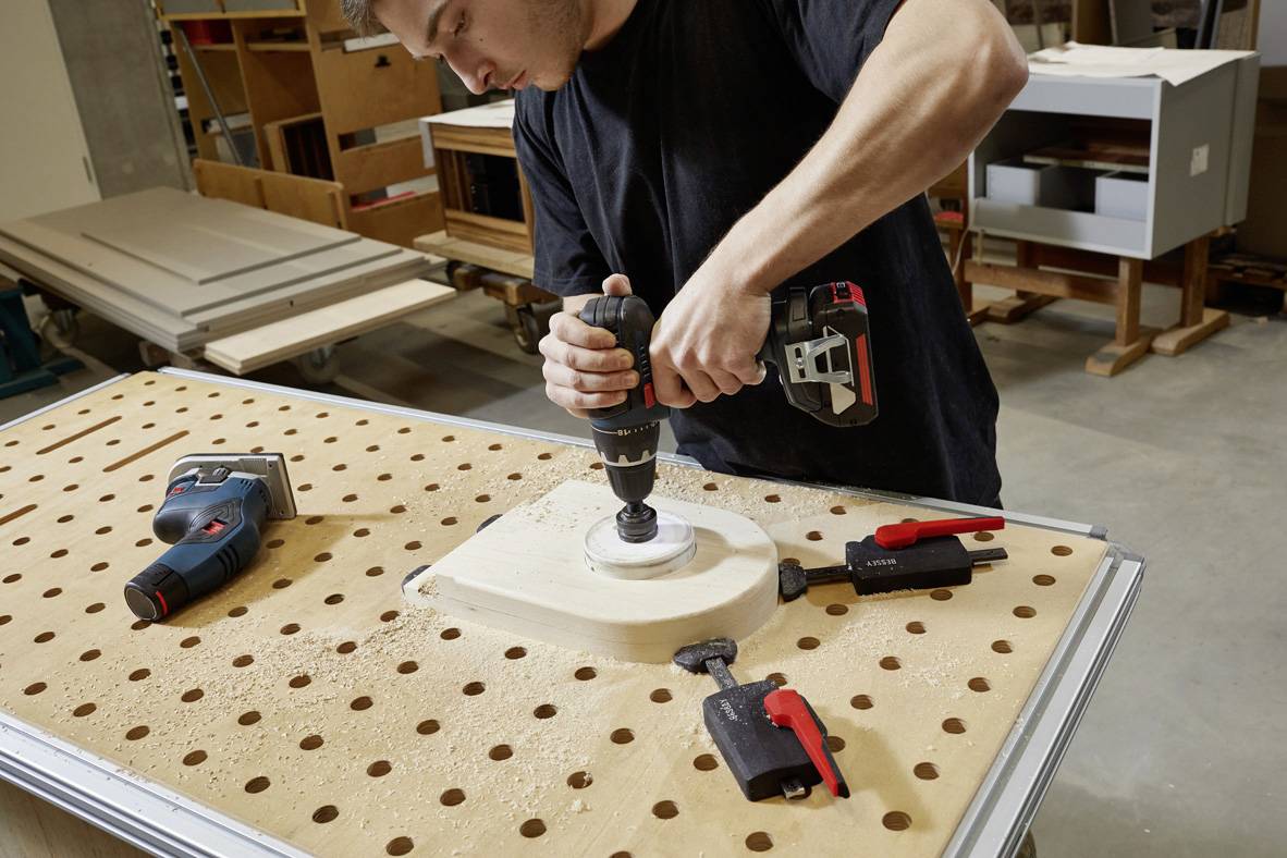 Ein Mann in einer Werkstatt bohrt ein rundes Loch in ein Holzstück, das auf einem Arbeitstisch mit Schraubzwingen befestigt ist.