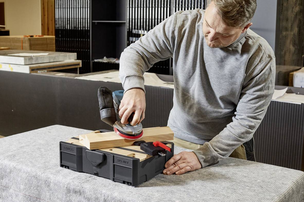 Ein Mann schleift ein Holzstück mit einem Elektroschleifer, während es mit einer Zwinge auf einer Werkbank befestigt ist.