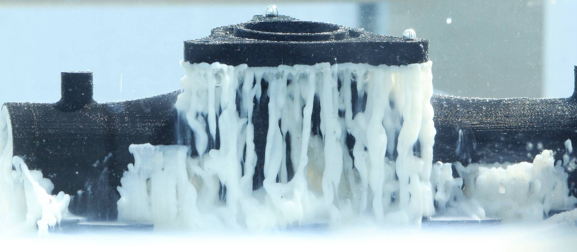 Ein 3D-Druckobjekt mit Stützkonstruktion wird von überschüssigem Material befreit. Weißes Material tropft von der schwarzen Struktur.