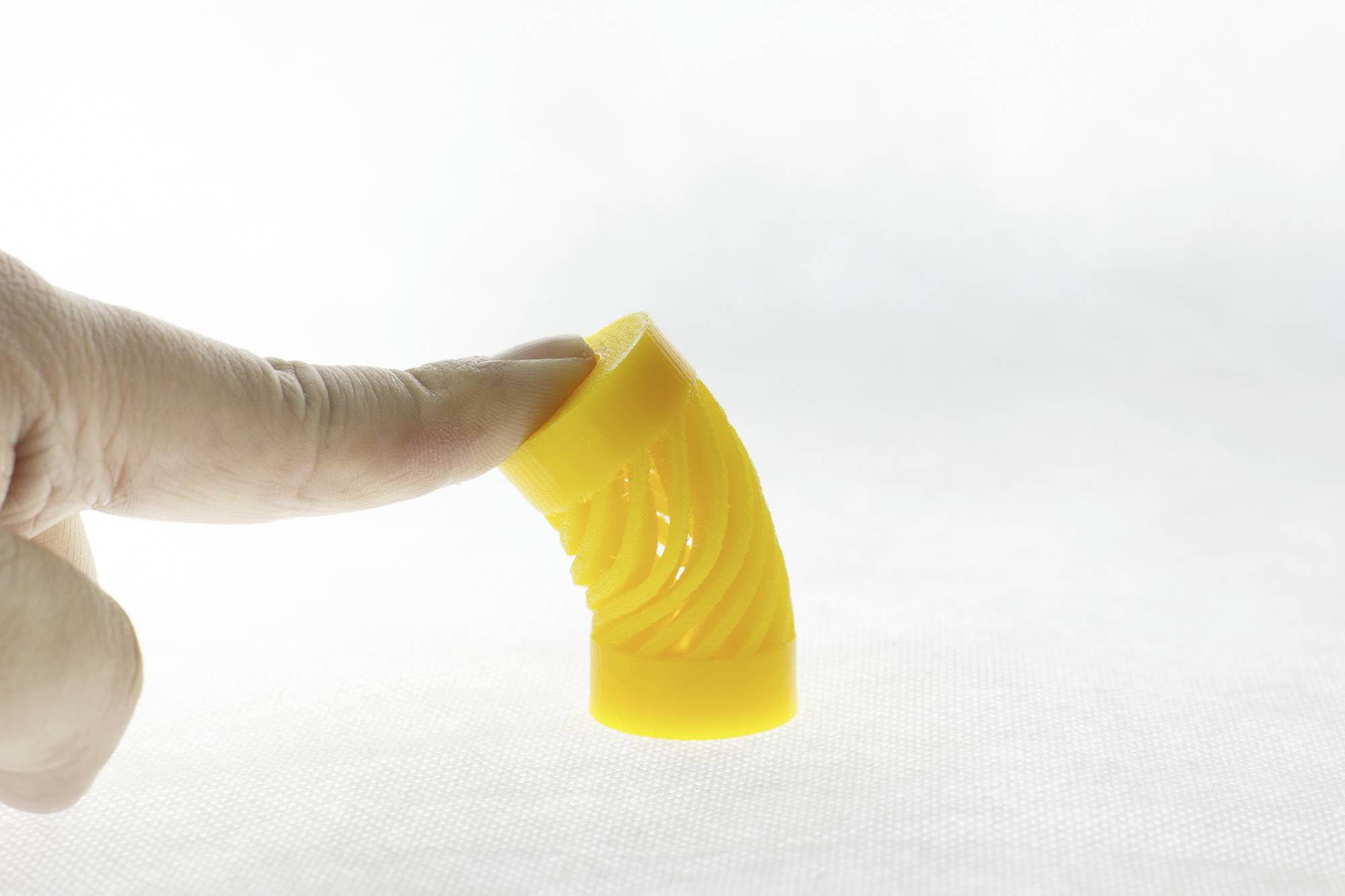 Eine Hand berührt ein flexibles, gelbes Kunststoffteil, das gebogen ist. Das Bild zeigt die Elastizität des Materials.