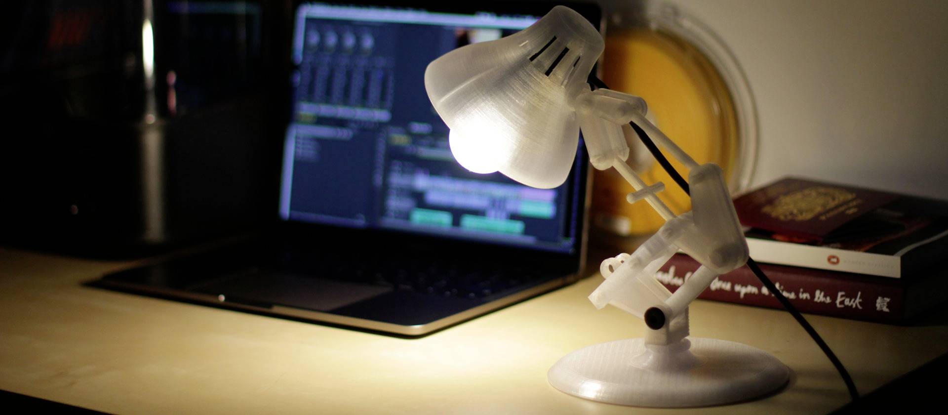 Ein Schreibtisch mit einer beleuchteten Lampe, einem Laptop mit geöffnetem Bildbearbeitungsprogramm und Büchern im Hintergrund.