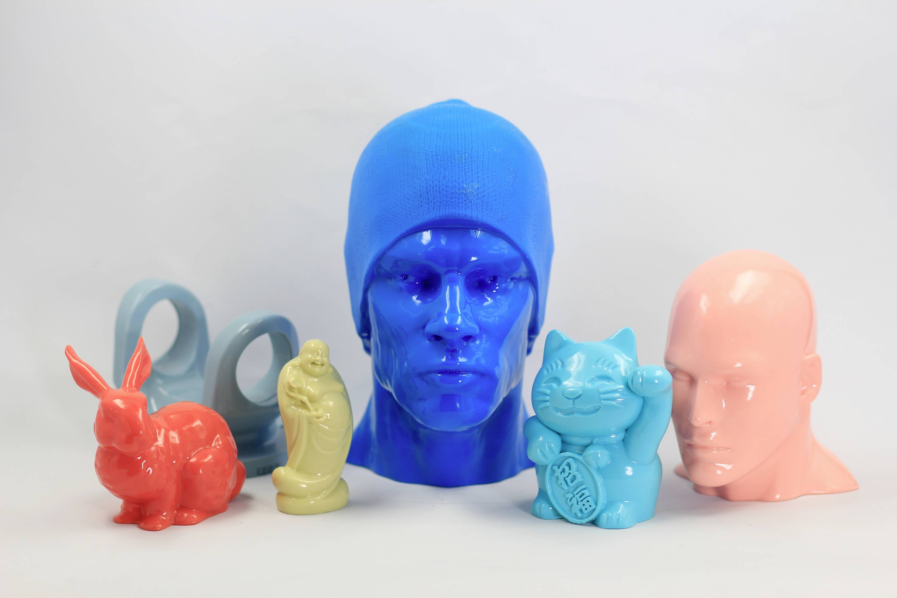 Bunte 3D-gedruckte Figuren: ein blauer Kopf mit Mütze, ein pinker Kopf, ein roter Hase, eine beige Figur, ein lachender blauer Katze und zwei blaue Ringe.