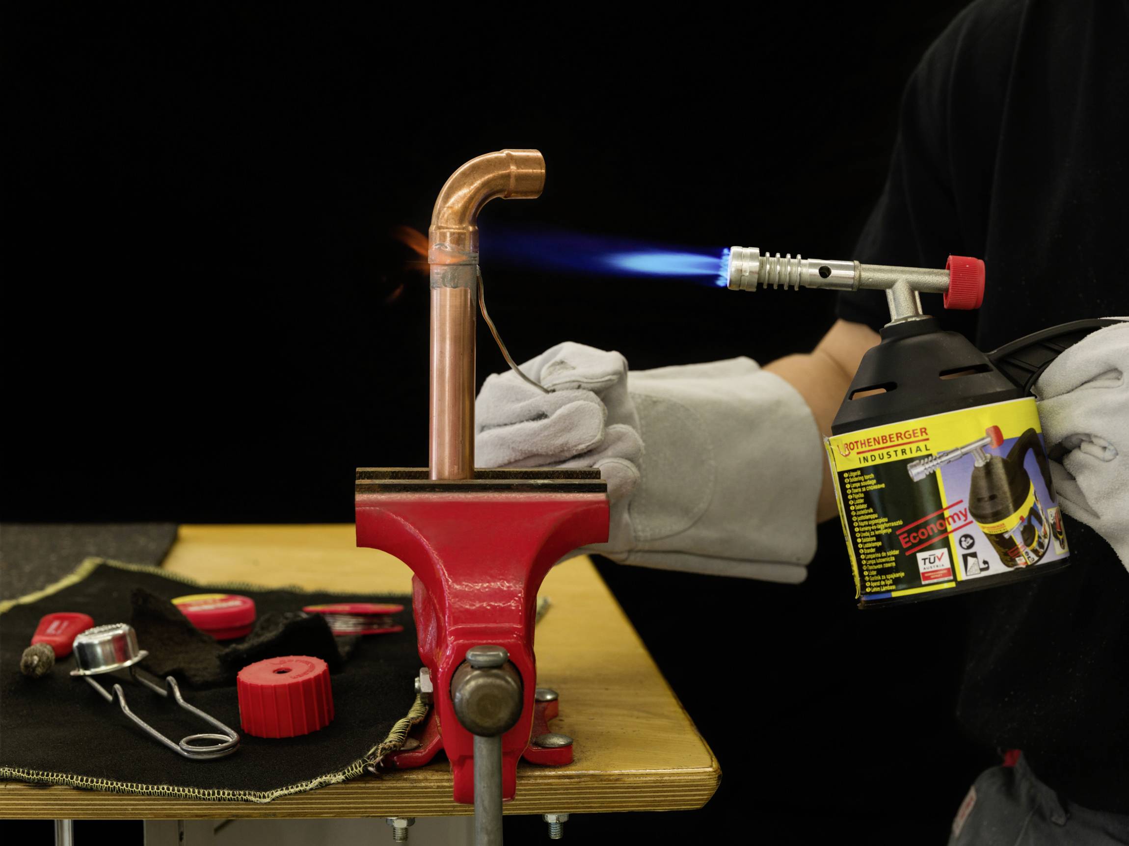 Eine Person lötet ein Kupferrohr mit einem Brenner. Das Rohr ist in einem Schraubstock fixiert. Schutzkleidung wird getragen.