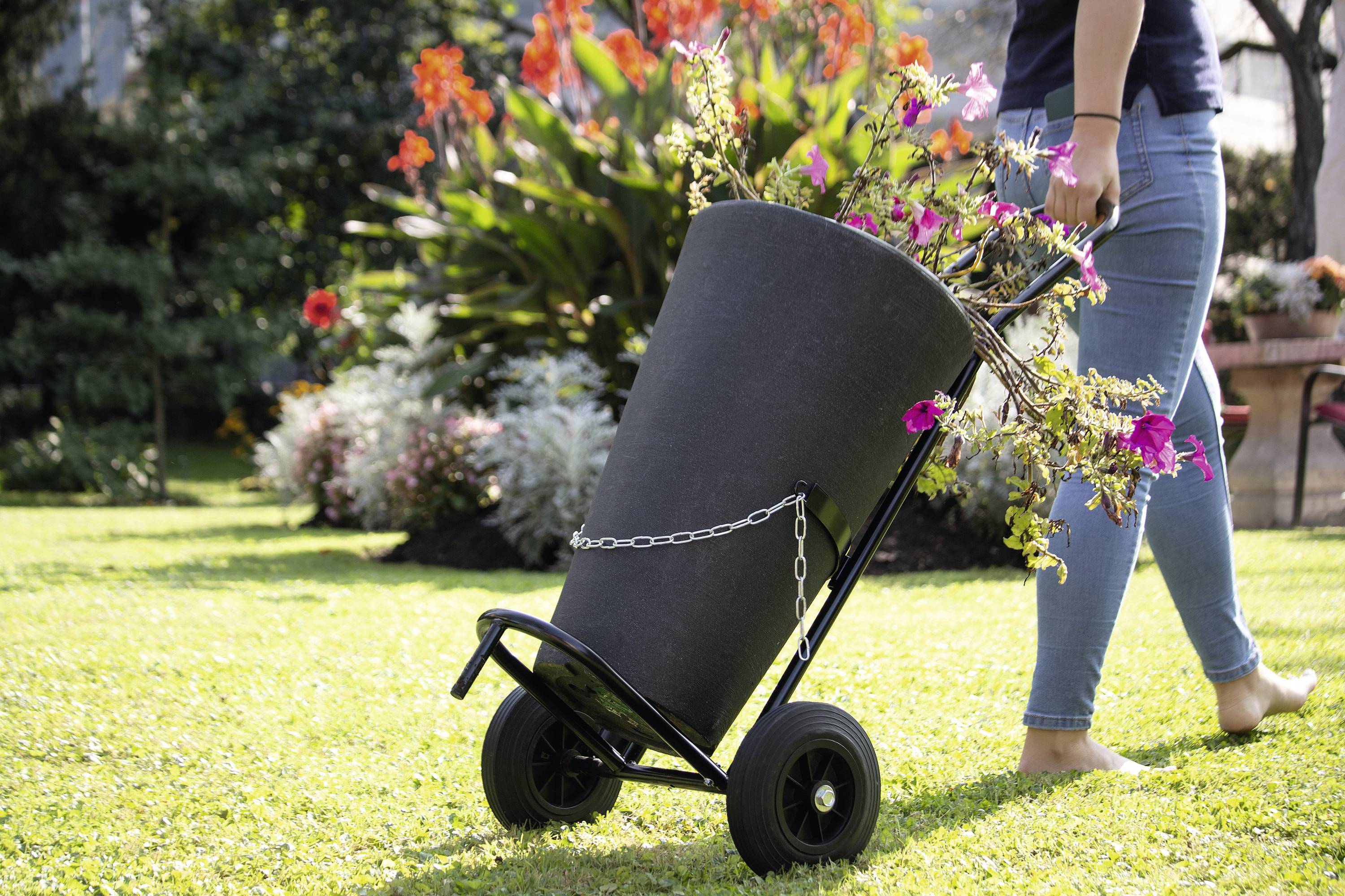 Eine Person transportiert einen großen Korb mit Blumen auf einem Handkarren über eine grüne Rasenfläche im Garten.