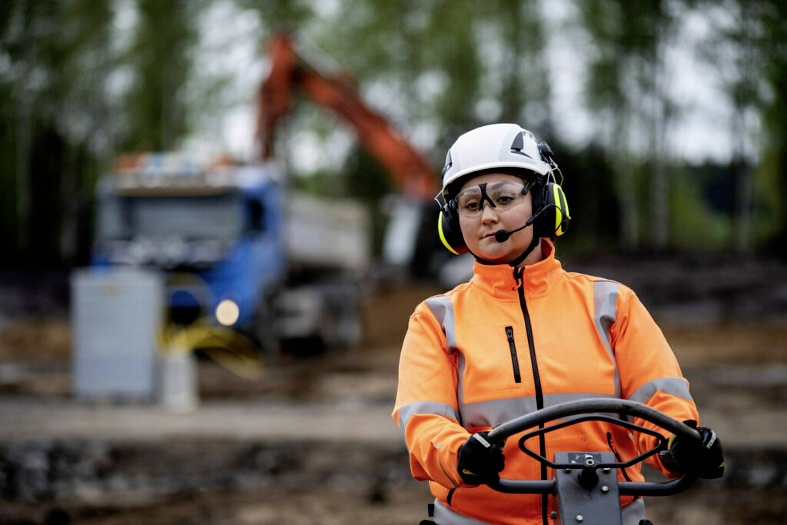 Eine Person in orangefarbener Schutzkleidung und Helm bedient schwere Maschinen auf einer Baustelle; im Hintergrund unscharf ein Lkw.