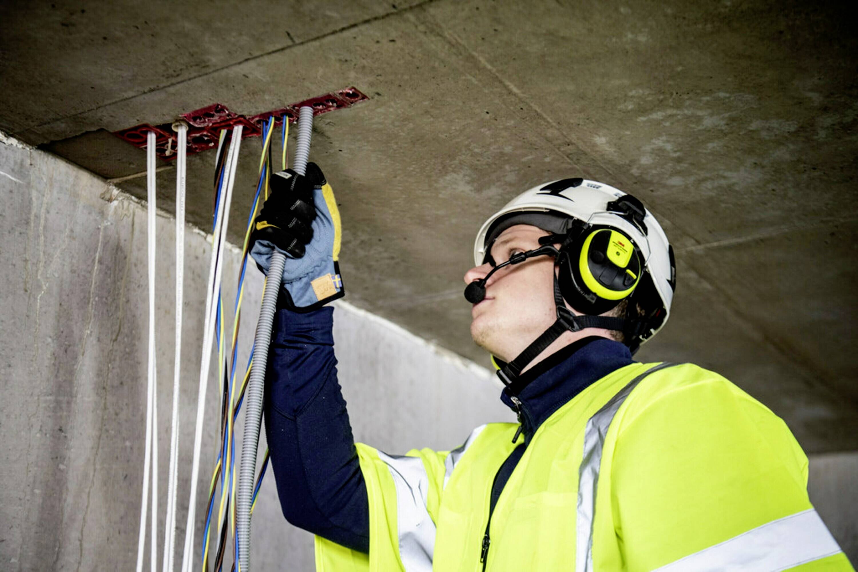 Ein Arbeiter mit Schutzhelm und Warnweste überprüft Kabel, die aus der Decke kommen. Er trägt Handschuhe und Gehörschutz.
