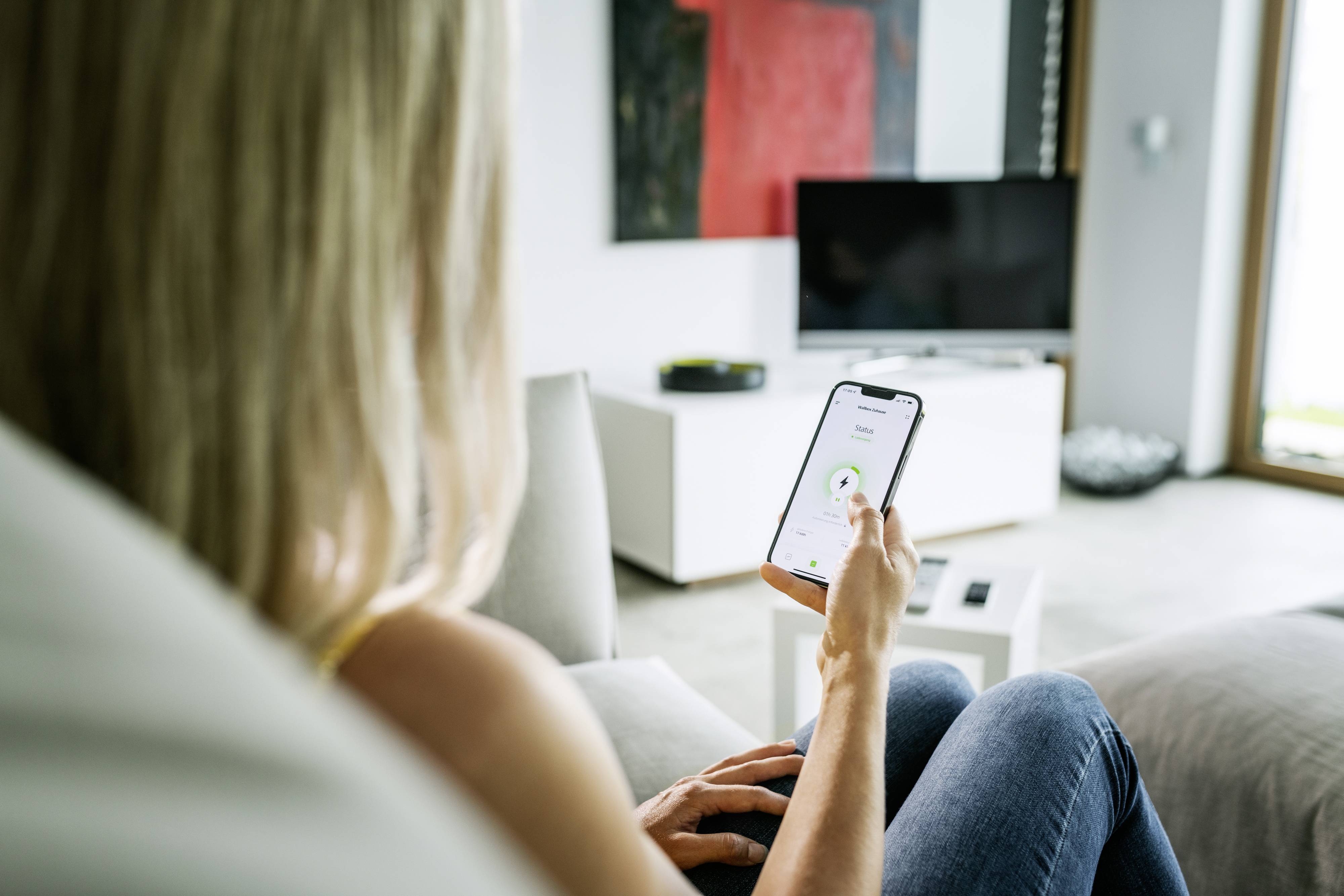 Eine Person sitzt auf einem Sofa und steuert ein Gerät mit dem Smartphone, auf dem ein grüner Button sichtbar ist, in einem modernen Wohnzimmer.