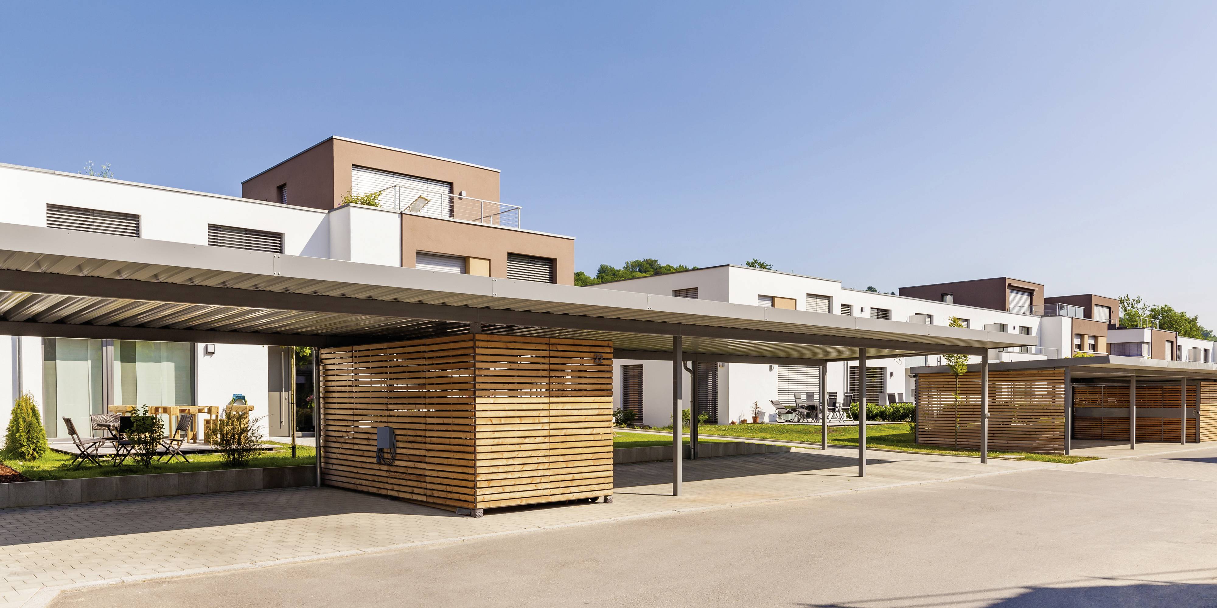 Moderne Reihenhäuser mit Flachdächern und Carports. Im Vordergrund sind die Carports aus Holzlamellen zu sehen. Klarer Himmel.