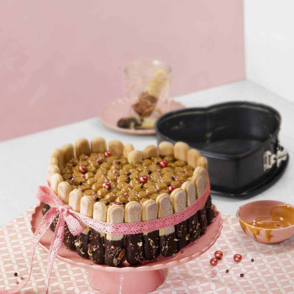 Herzförmiger Kuchen mit Keksrand, mit rosa Band umwickelt, dekoriert mit roten Perlen. Im Hintergrund eine leere Herzform.