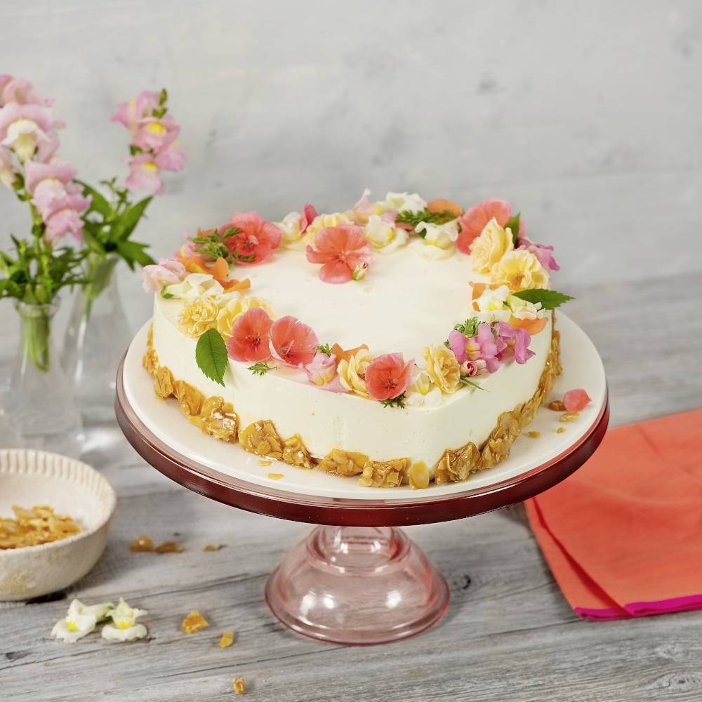 Eine runde Torte auf einem rosa Kuchenständer, dekoriert mit bunten essbaren Blumen, steht auf einem Tisch neben einer Vase mit Blüten.